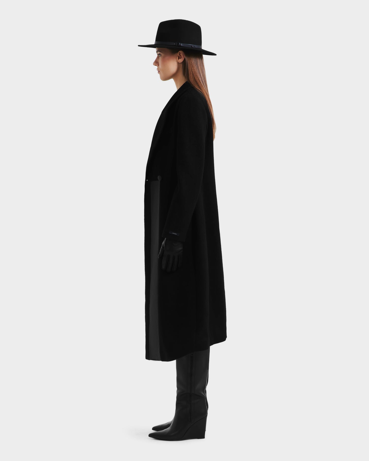 A woman in the Rudsak LYNWOOD - 8125539 Black premium wool coat with a leather front panel, black gloves, wide-brim hat, and knee-high boots stands in profile against a plain light background.