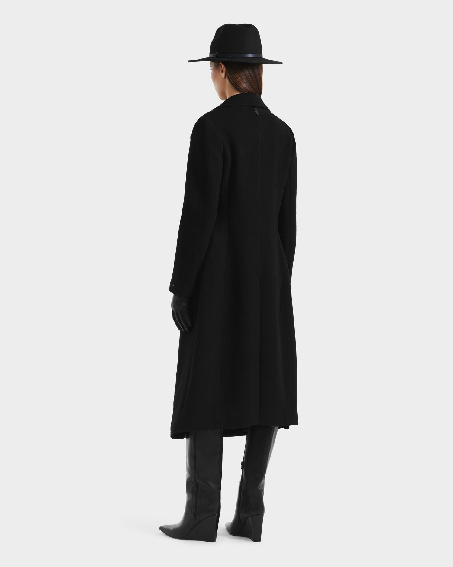 A person faces away, wearing the Rudsak LYNWOOD - 8125539 Black womens long tailored coat with a leather front panel, paired with black gloves, knee-high boots, and a wide-brimmed hat against a plain light background.