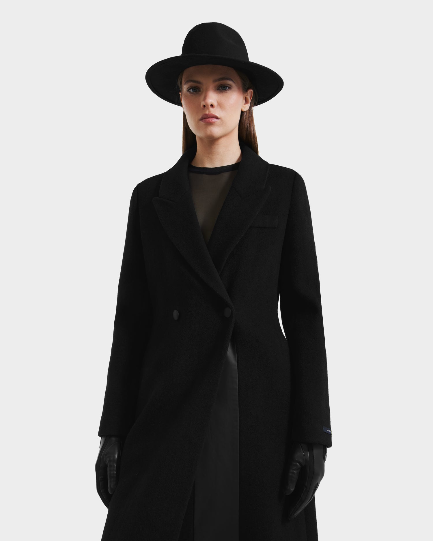 A woman in the Rudsak LYNWOOD - 8125539 Black long tailored coat stands against a white background, wearing a wide-brimmed hat, black gloves, and a sheer black top, exuding effortless sophistication with her neutral expression.