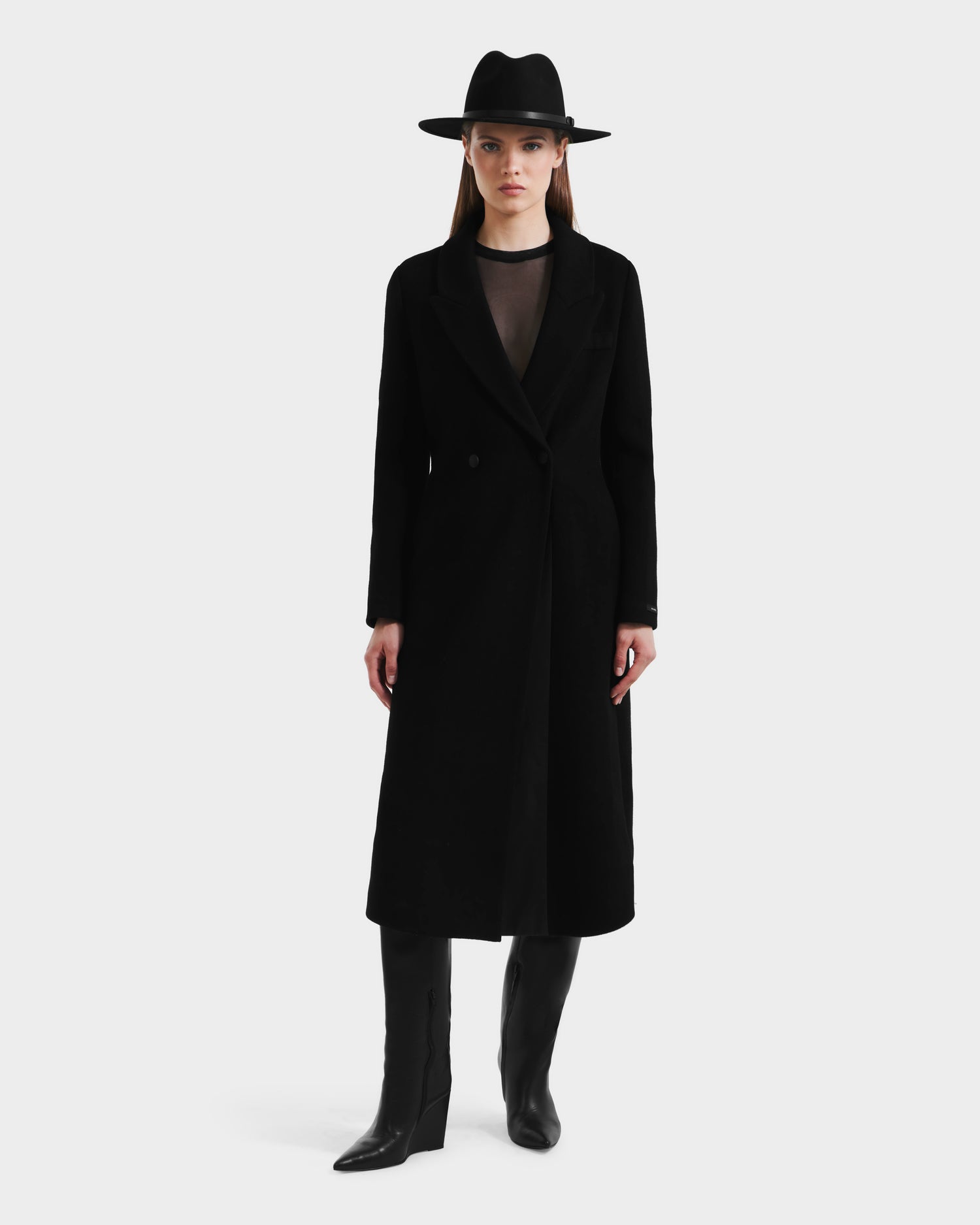 A woman faces forward in the Rudsak LYNWOOD - 8125539 Black long tailored coat with a leather front panel, paired with black knee-high boots and a wide-brimmed hat, her straight brown hair framing her neutral expression against a plain light background.