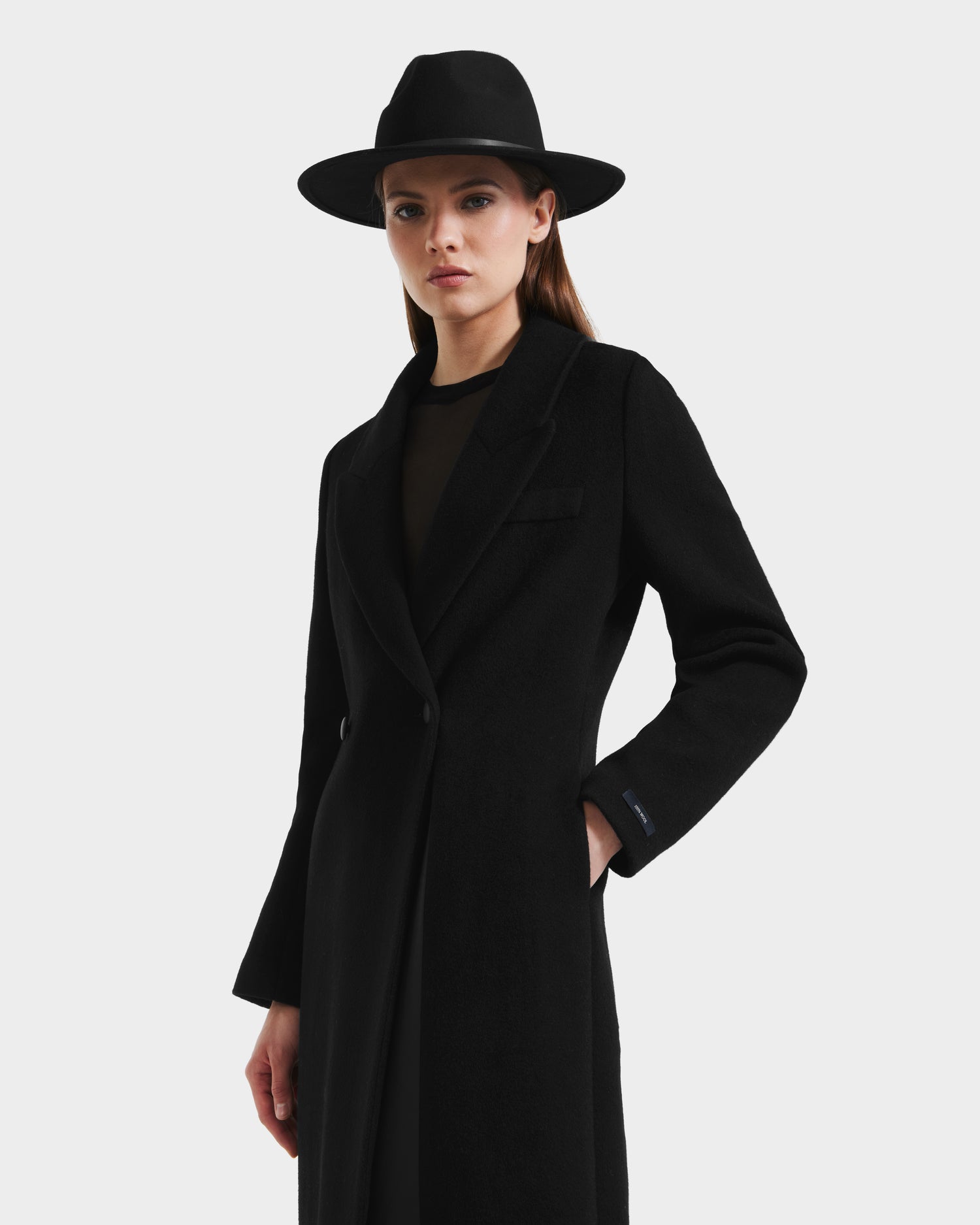 A woman wearing the Rudsak LYNWOOD - 8125539 Black tailored coat and a wide-brimmed black hat stands with one hand in her pocket against a plain white background.