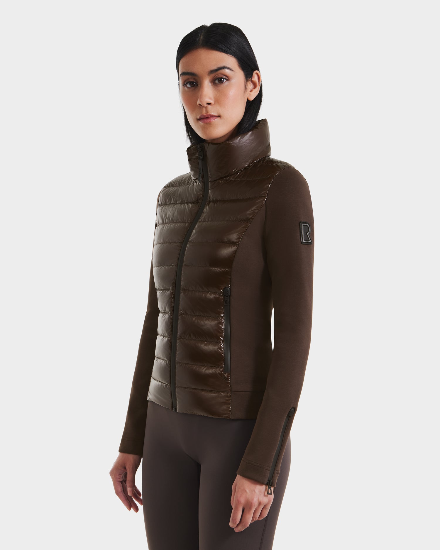 A woman with dark hair models the Rudsak MABS C - 8124590 Espresso, a fitted brown lightweight activewear jacket with a high collar and dark brown sleeves, standing facing forward against a plain white background.