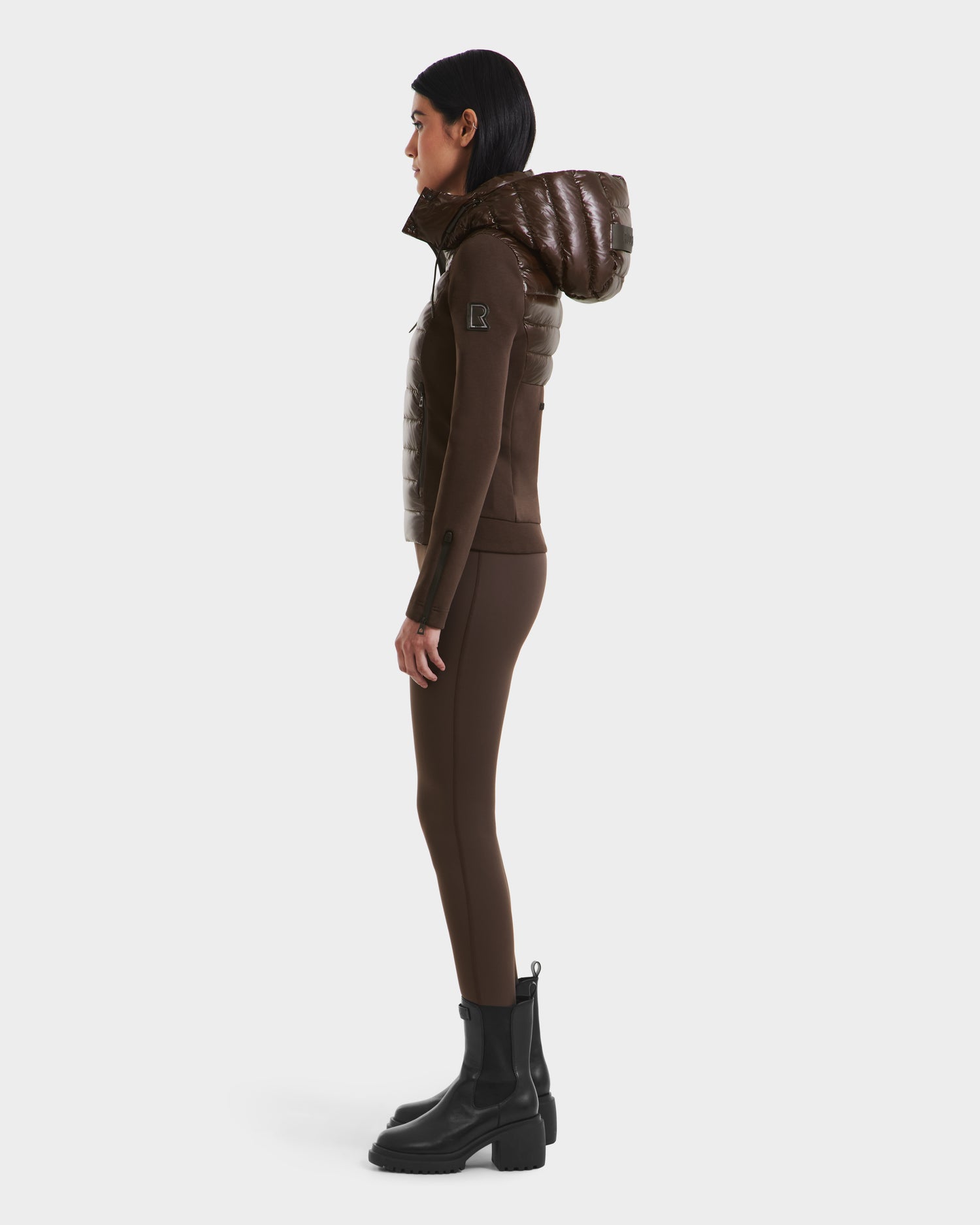 A woman stands in profile wearing the Rudsak MABS C - 8124590 Espresso shiny brown puffer jacket with a hood, tight brown pants, and black ankle boots against a plain white background.