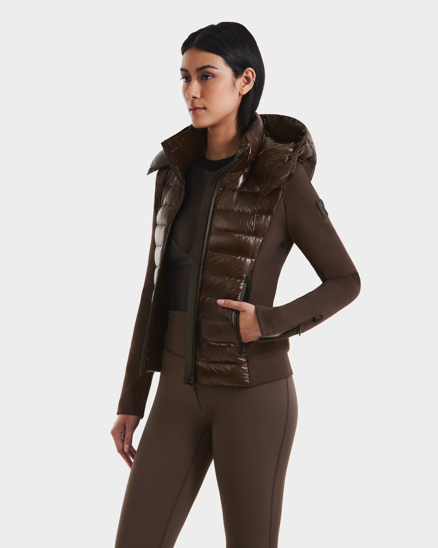 A woman wearing the Rudsak MABS C - 8124590 Espresso, a brown fitted puffer jacket with glossy quilted panels and matching brown leggings, stands against a plain white background.