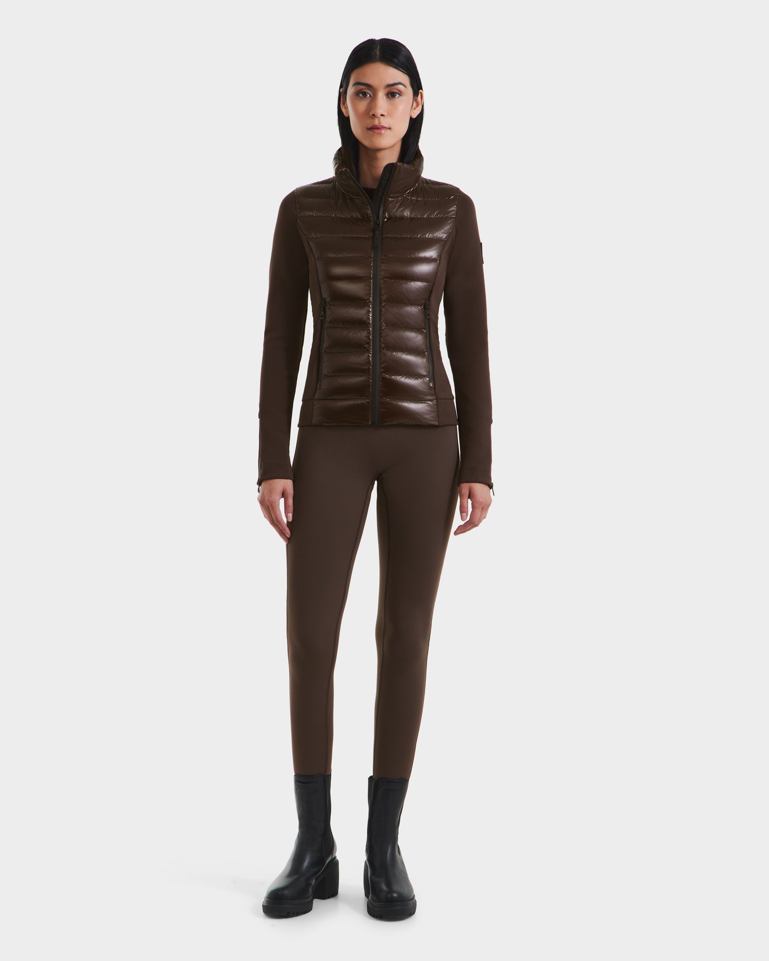 A woman stands facing forward wearing the Rudsak MABS C - 8124590 Espresso, a fitted, shiny brown puffer jacket filled with RDS-certified down, paired with matching brown leggings and black ankle boots against a white background.