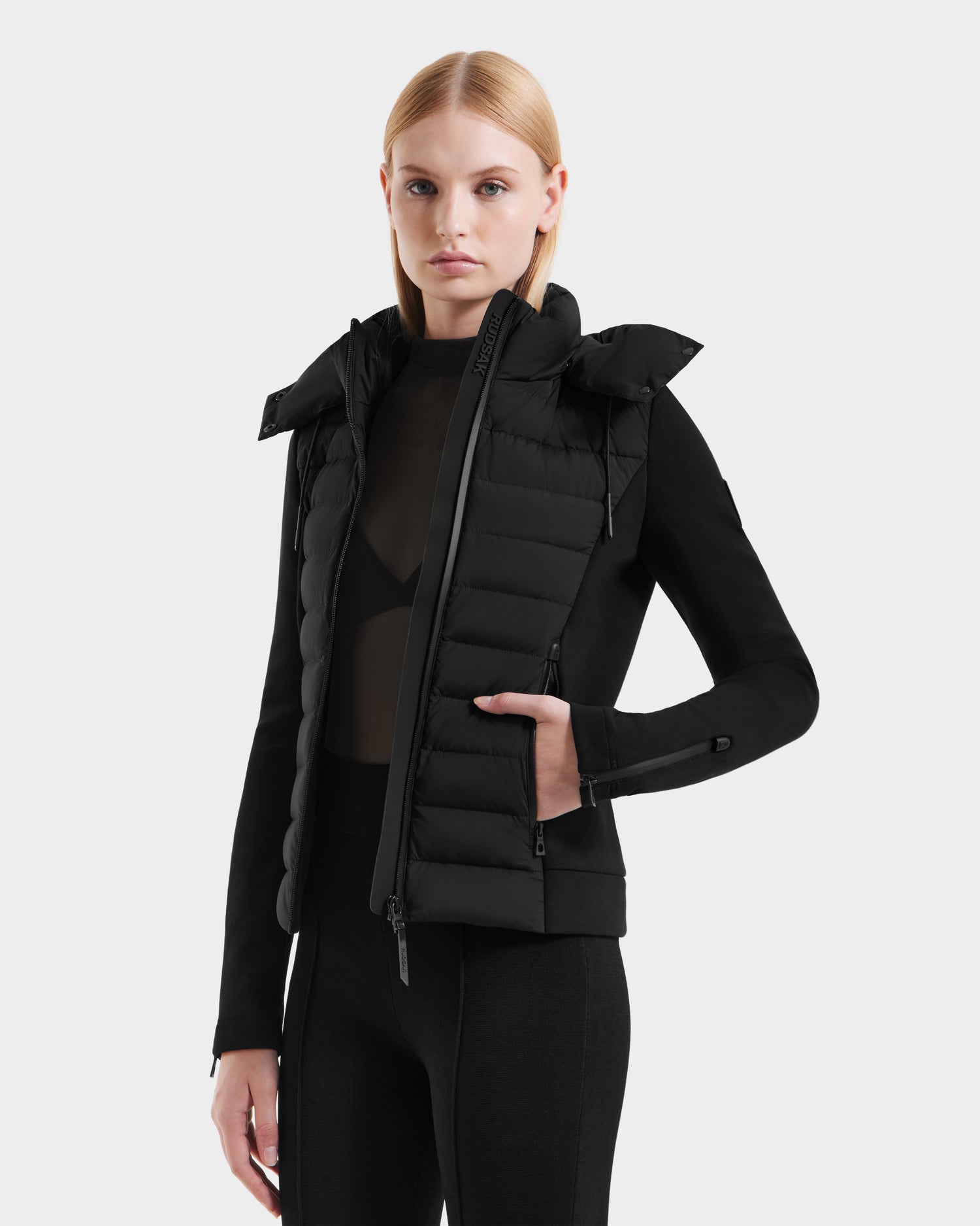 A woman with straight blonde hair models the Rudsak MABS - 8125055 Black fitted women’s puffer jacket with a high collar over a sheer black top and black pants, standing against a plain white background.