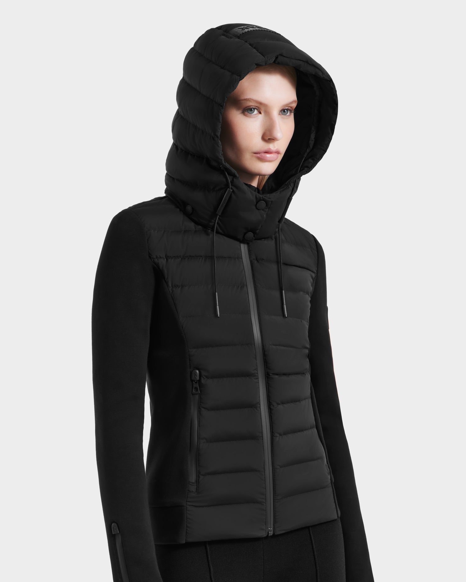 A woman wears the Rudsak MABS - 8125055 Black quilted hooded womens puffer jacket with zippered pockets, standing against a plain white background.