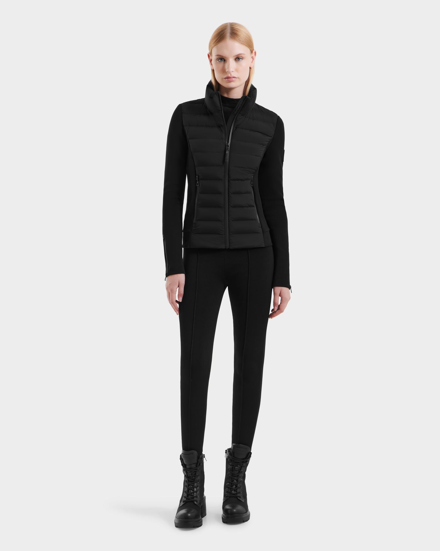 A woman with straight blond hair faces forward, wearing the Rudsak MABS - 8125055 Black womens puffer jacket, black leggings, and lace-up boots against a plain white background.