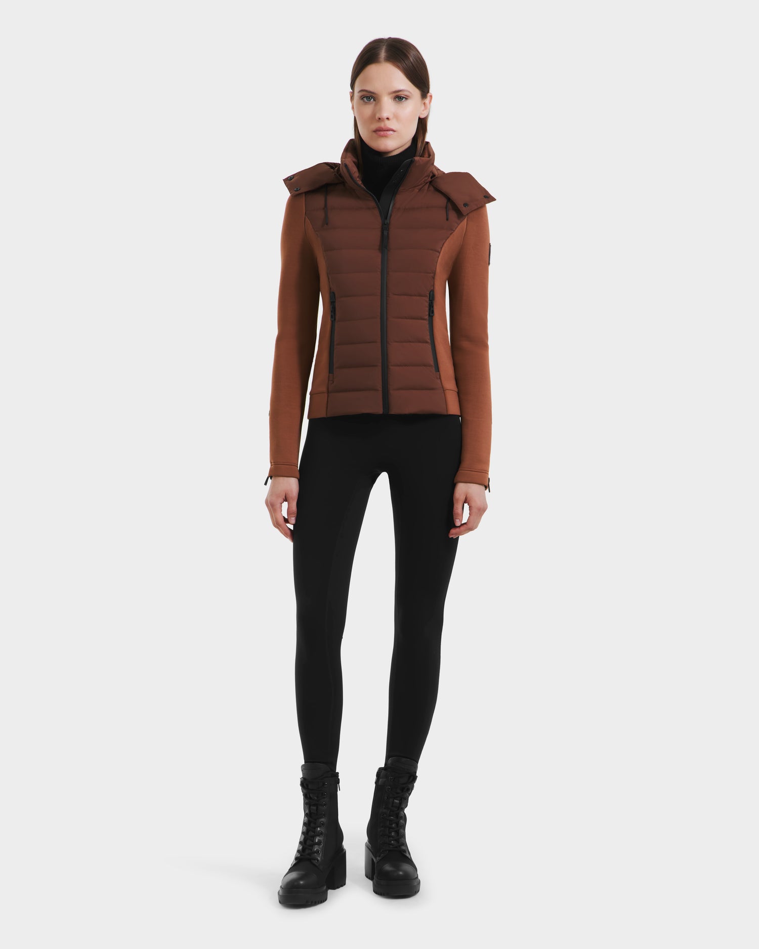 A woman with straight hair stands facing forward against a white background, wearing the Rudsak MABS - 8125055 Rust women’s puffer jacket with hood. This RDS-certified down brown jacket pairs with black leggings and lace-up boots for warmth and style.