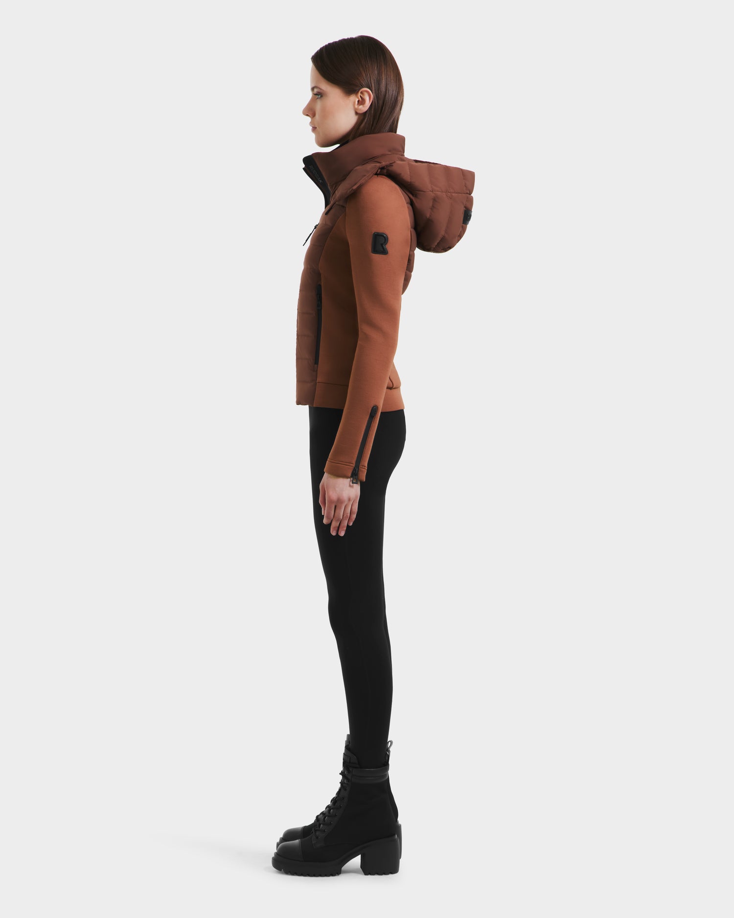 A woman in profile wears the Rudsak MABS - 8125055 Rust women’s puffer jacket with black leggings and lace-up boots, standing against a plain white background.
