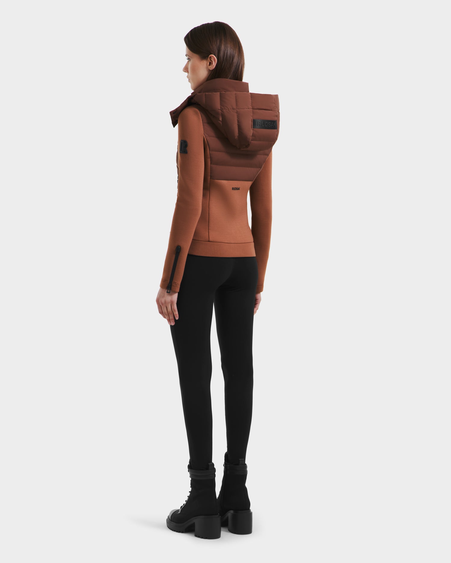 A woman stands facing away, wearing the Rudsak MABS - 8125055 Rust womens puffer jacket with a large hood, paired with black leggings and heeled boots against a plain white background.