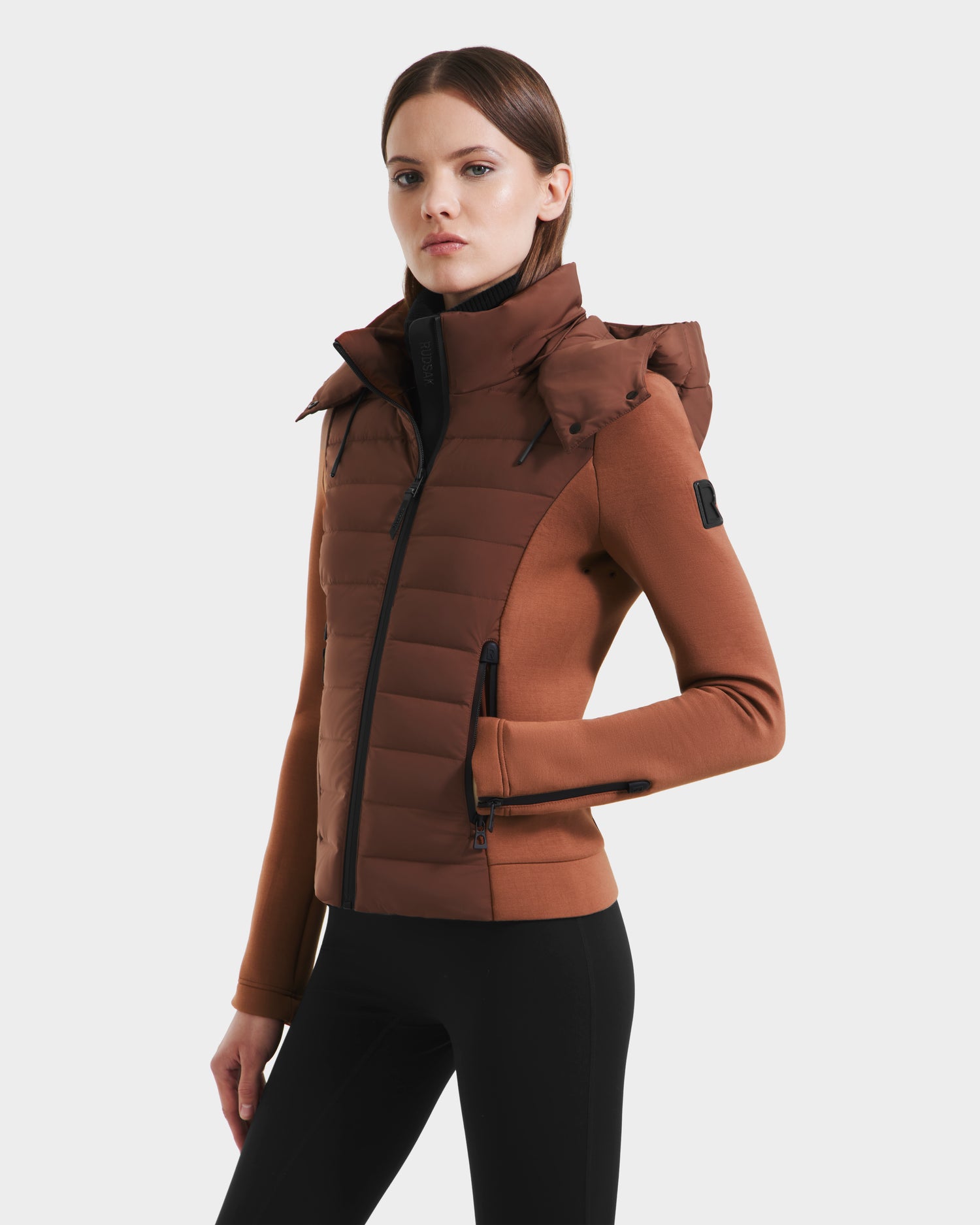 A woman wearing the Rudsak MABS - 8125055 Rust womens puffer jacket with a high collar and hood, paired with black pants, stands against a white background. She has straight brown hair and a neutral expression.