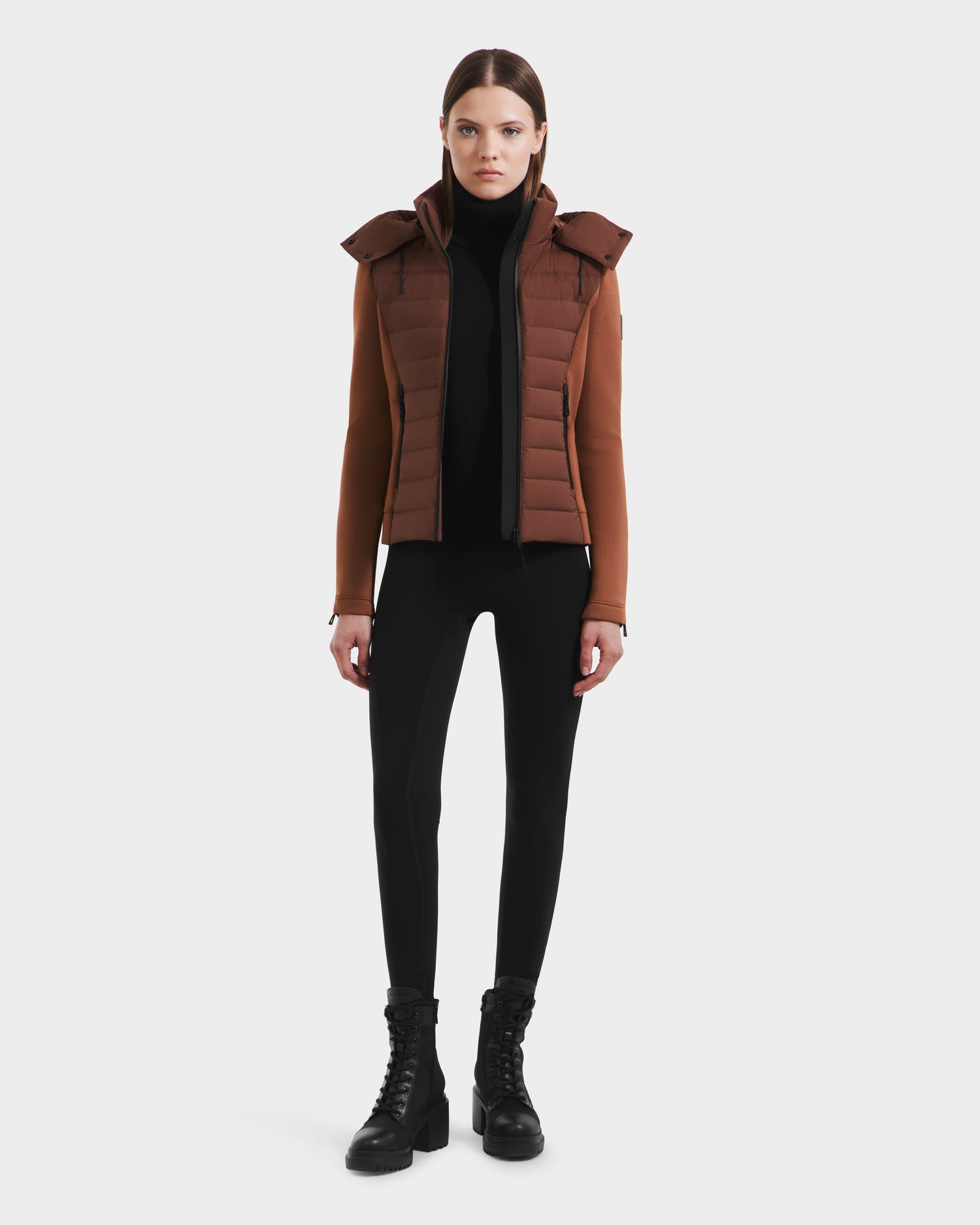 A woman faces forward against a white background, wearing the Rudsak MABS - 8125055 Rust women’s puffer jacket with a hood, paired with a black turtleneck, black leggings, and black lace-up boots.