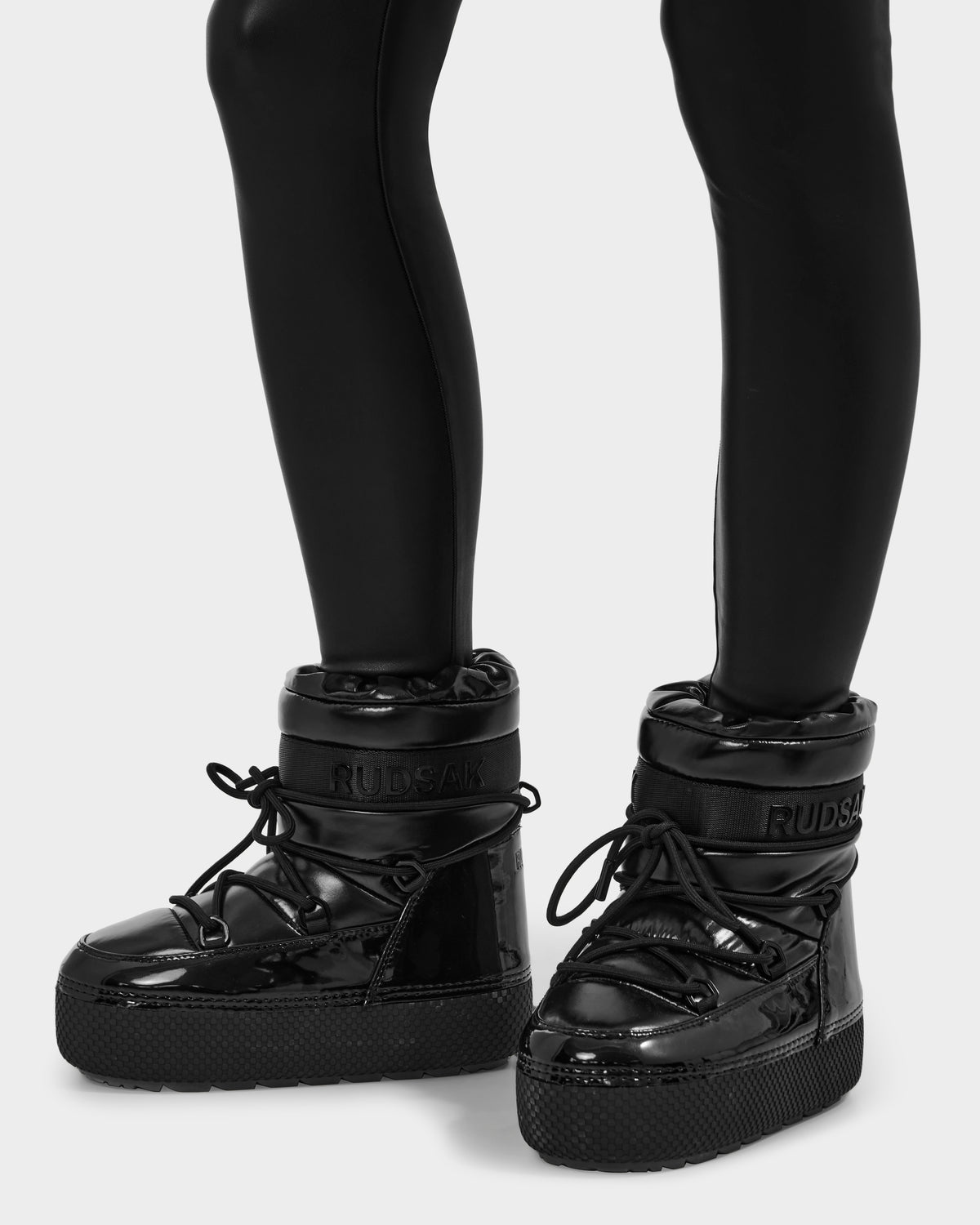 Person wearing shiny black Rudsak MACE - 8223500 platform boots with thick soles, laces, and cozy faux fur lining, paired with black leggings. "RUDSAK" branding appears on the upper front. The background is plain white.