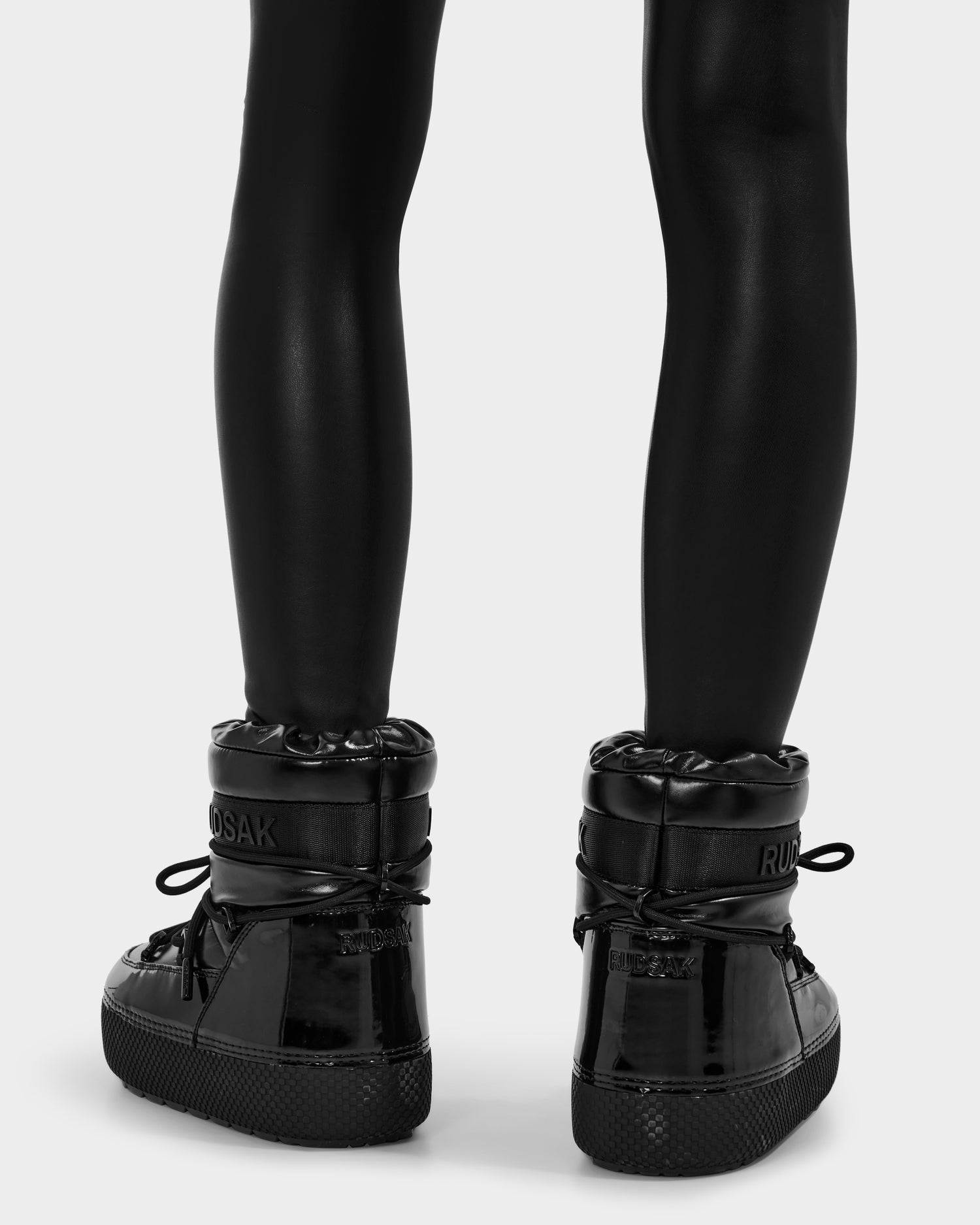 A person wears shiny black leggings and Rudsak MACE - 8223500 Black high-top sneakers, featuring thick soles, padded collars, and visible laces—evoking women’s moon boots. The background is plain and light-colored.