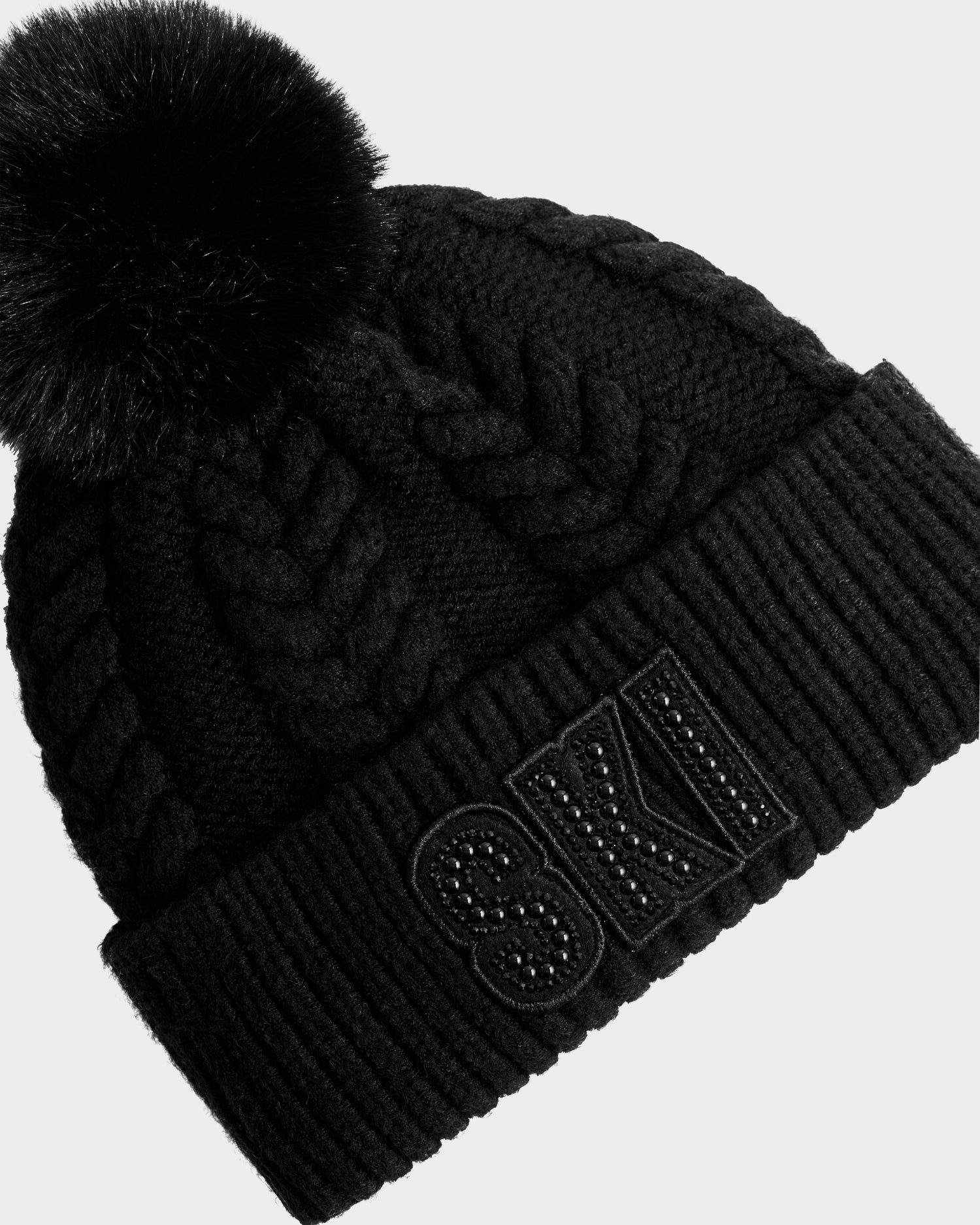 The Rudsak MAE - 8425550 Black is a black cable knit tuque with a chunky braided pattern, wide cuff featuring "SKI" in black studs, and a large faux fur pom pom for cozy slope-ready style.
