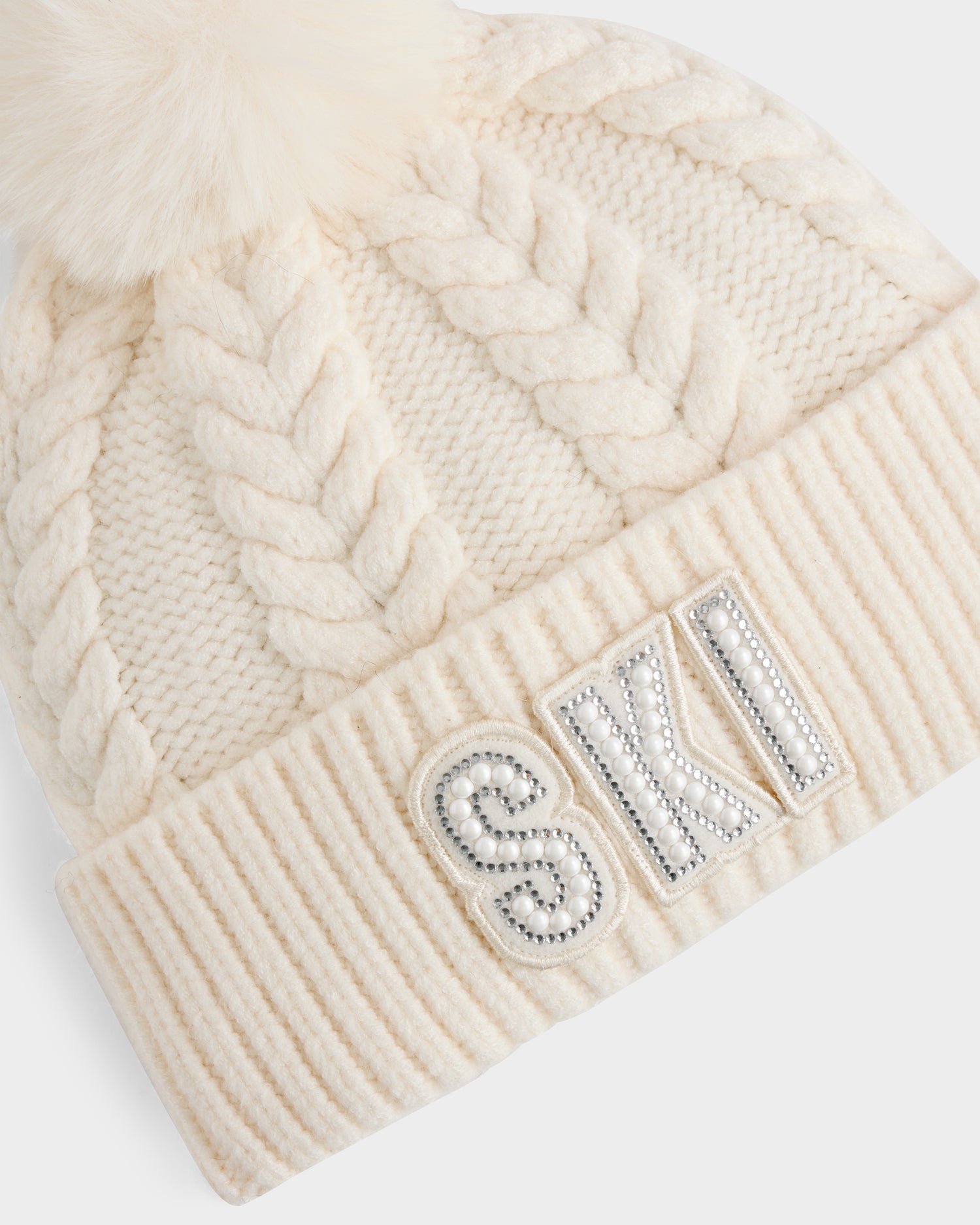 The MAE - 8425550 Vanilla by Rudsak is a cream women’s cable knit tuque featuring thick braided patterns, a faux fur pom pom, and a crystal-embellished SKI logo with rhinestones on the folded brim.