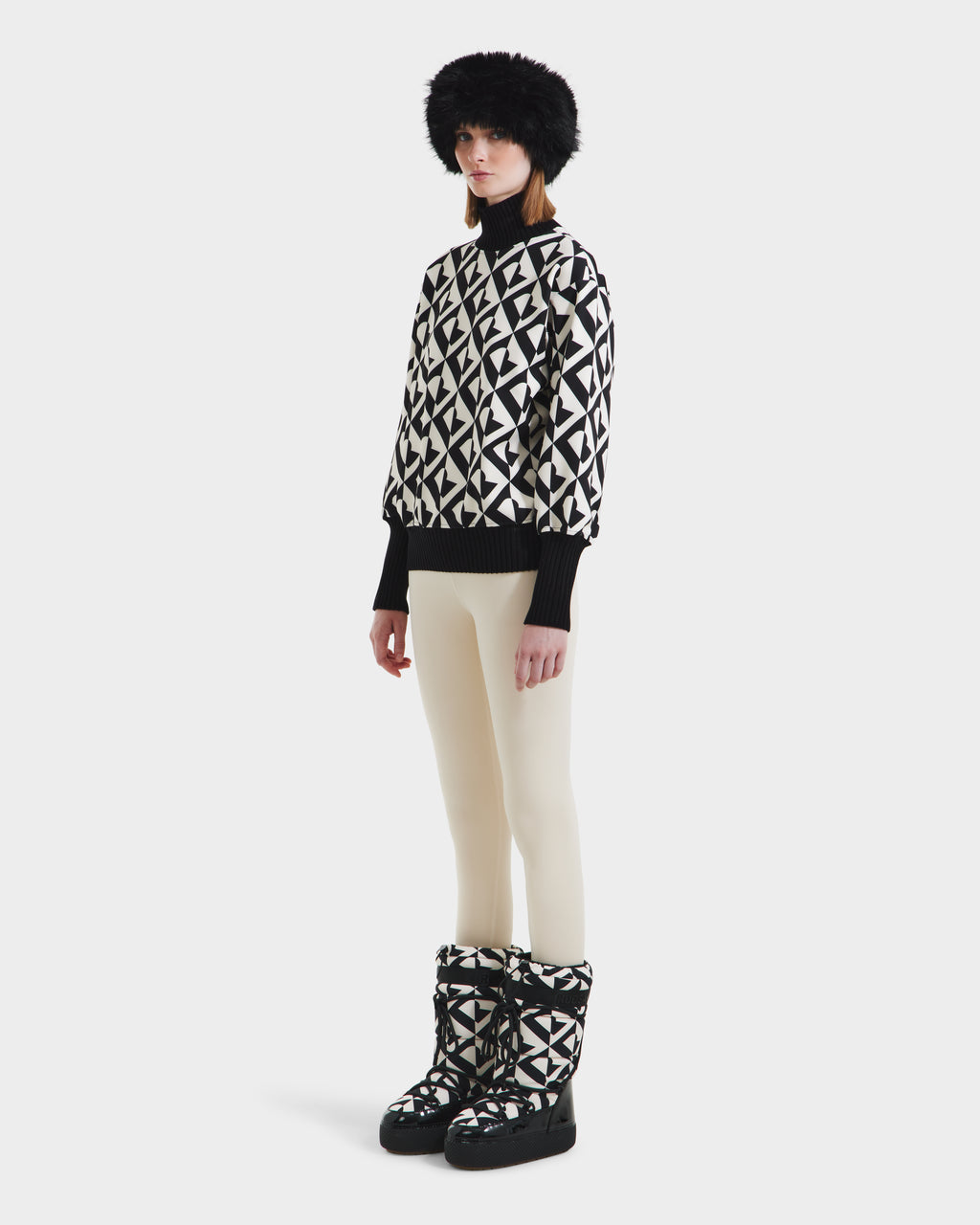 A woman stands against a plain white background wearing the Rudsak MAGGIE - 8525645 Vanilla Monogram ski sweater, styled with white pants, matching winter boots, black gloves, and a fluffy black hat.