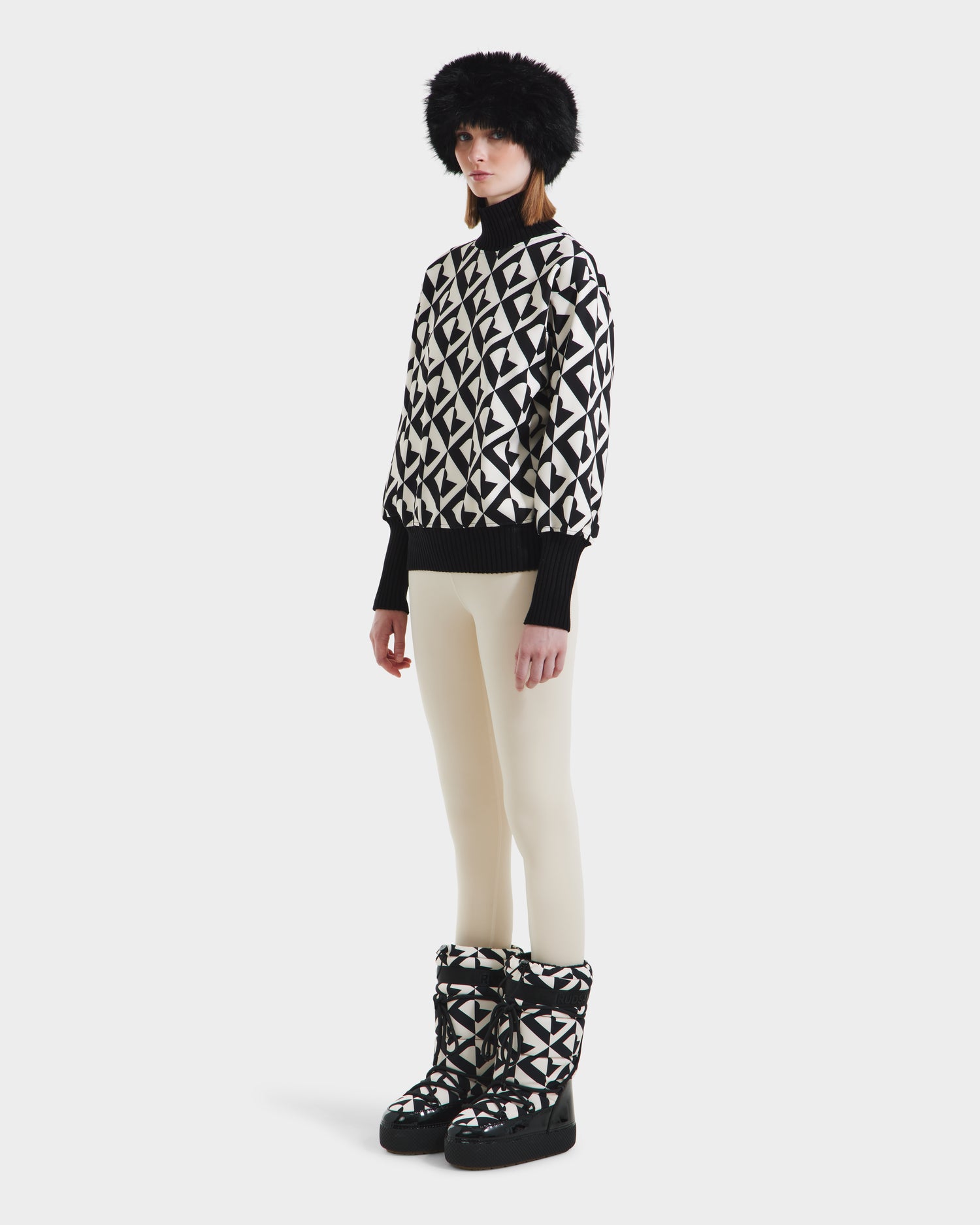 A woman stands against a plain white background wearing the Rudsak MAGGIE - 8525645 Vanilla Monogram ski sweater, styled with white pants, matching winter boots, black gloves, and a fluffy black hat.
