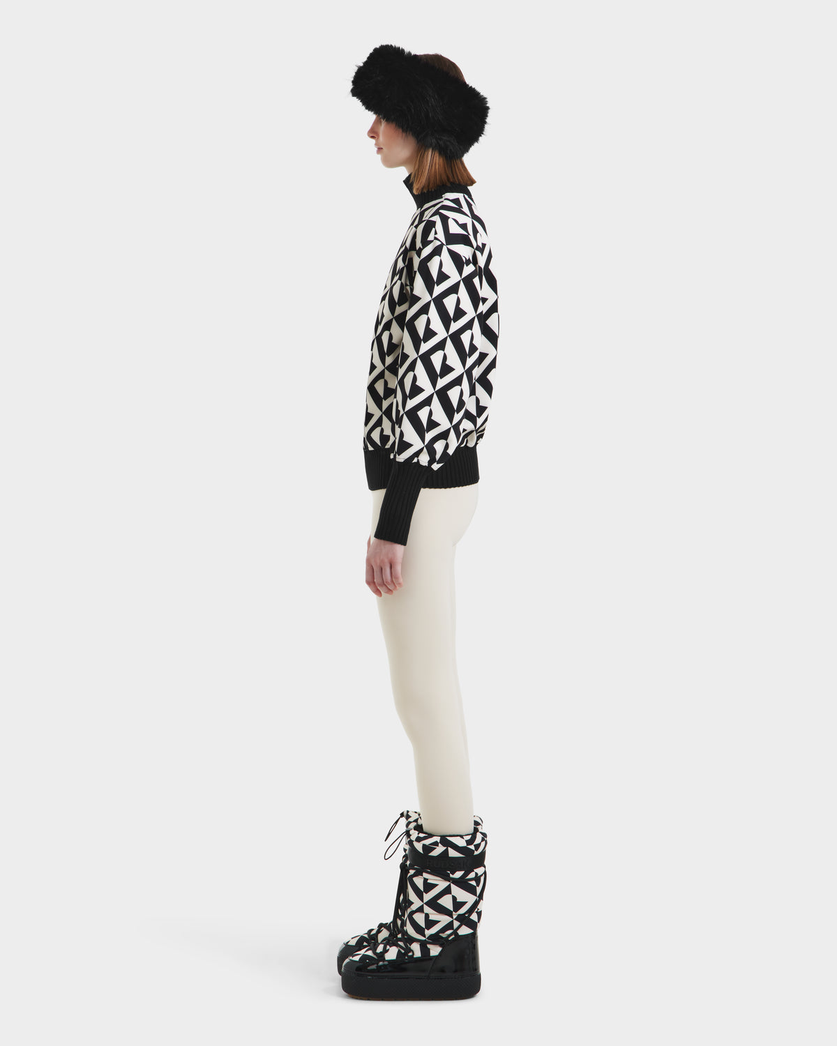 A woman stands in profile wearing Rudsak’s MAGGIE - 8525645 Vanilla Monogram winter boots, paired with a black fur visor, a geometric women’s ski sweater, and white pants against a plain white background.
