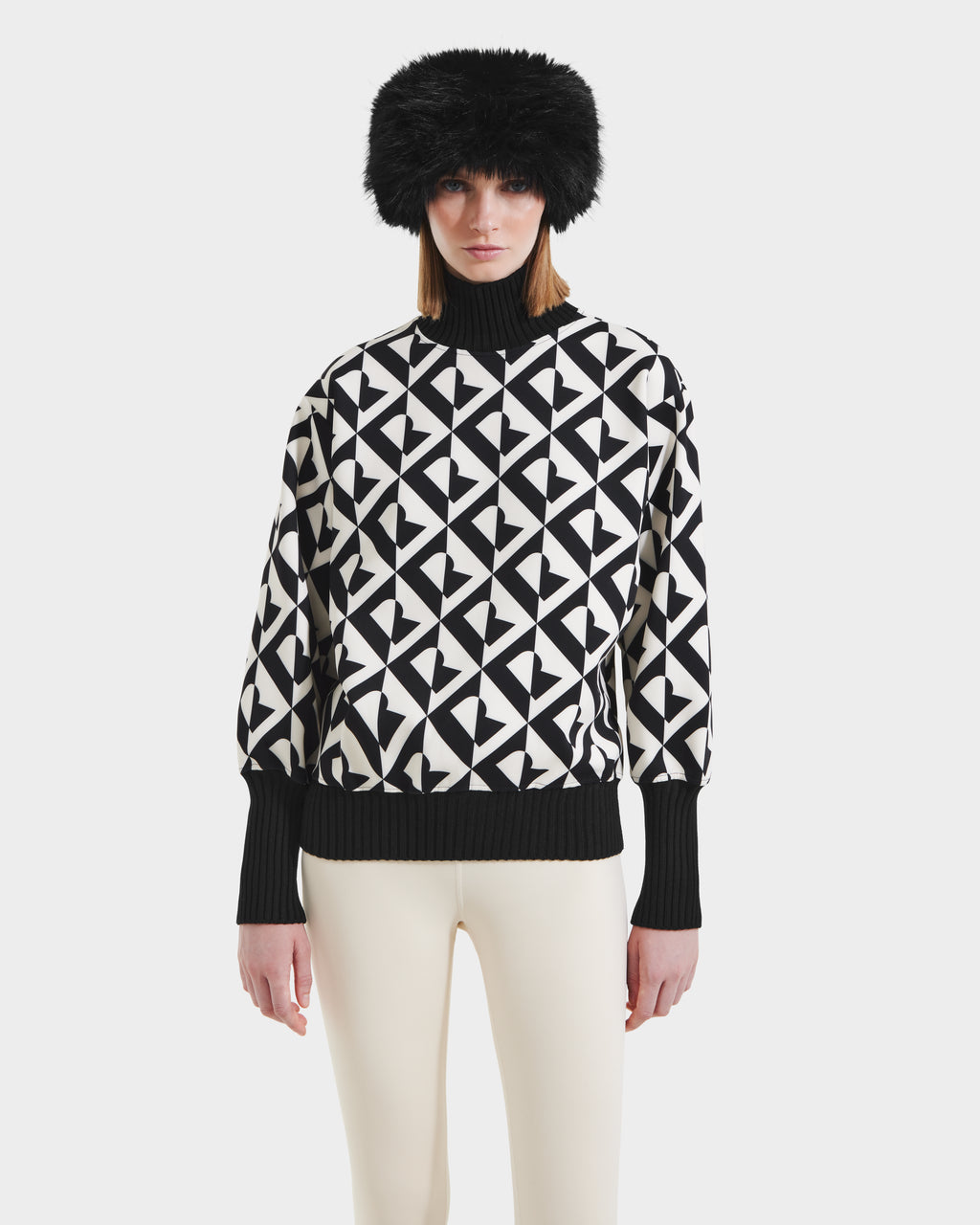 A woman models the Rudsak MAGGIE - 8525645 Vanilla Monogram ski sweater with black-and-white geometric patterns, paired with a black furry hat and white pants, standing against a plain white background.