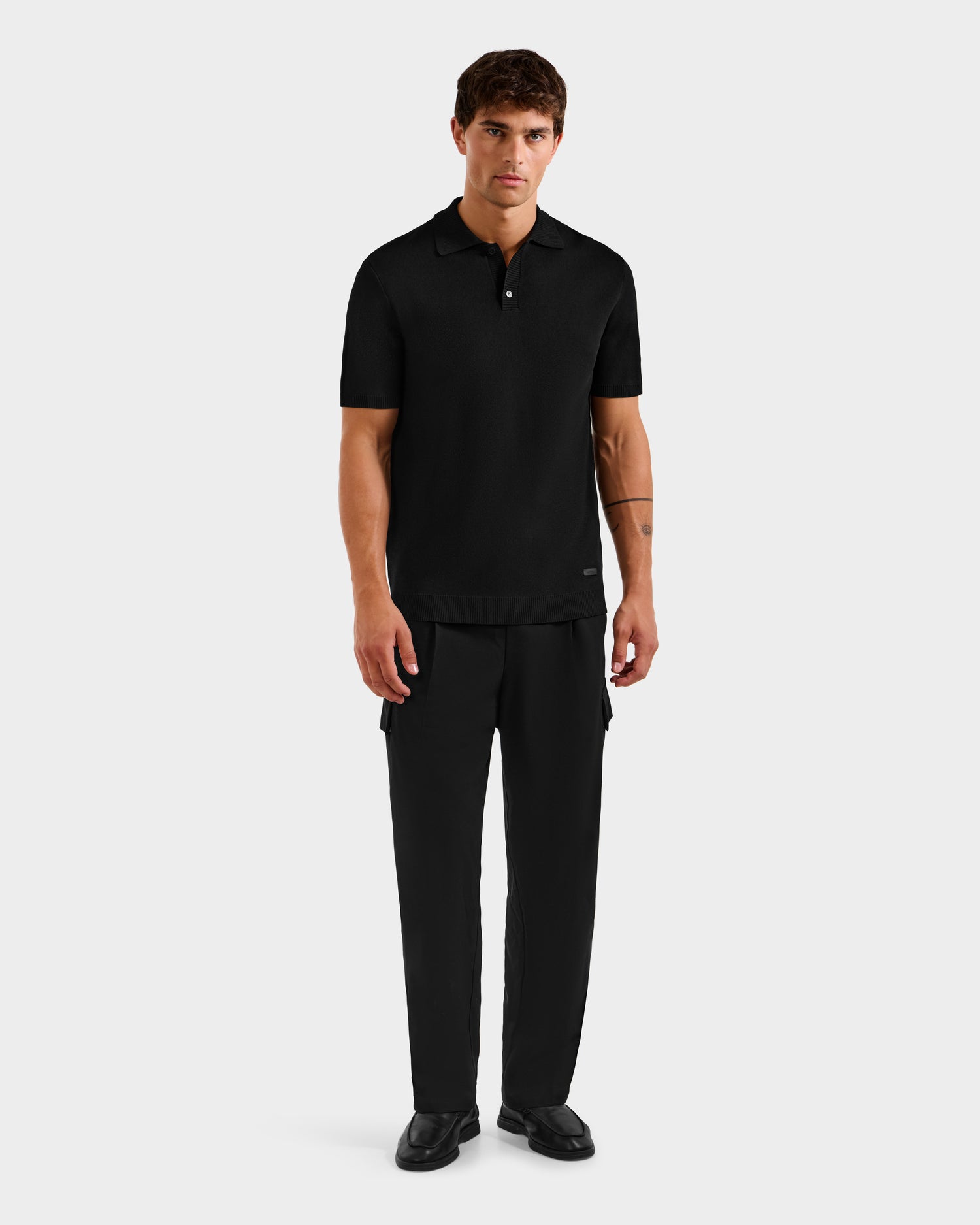A man with short brown hair and a neutral expression stands against a plain light grey background, wearing the Rudsak MAISEN - 6524518 Black polo, paired with black trousers and shoes. His arms are relaxed at his sides.