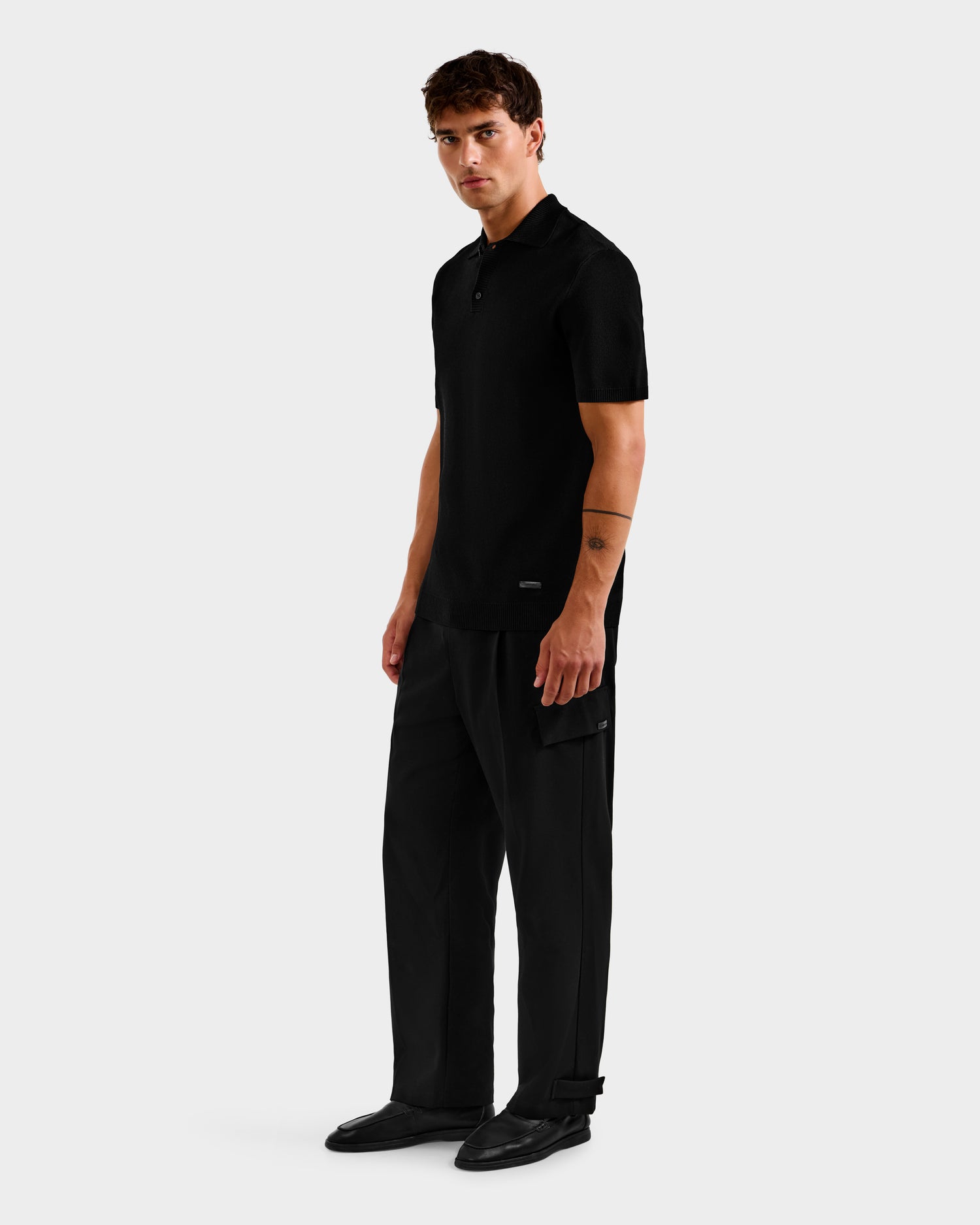 A man stands against a plain light gray background, wearing the MAISEN - 6524518 Black polo by Rudsak with wide-leg pants. He finishes the modern look with black shoes, short brown hair, and a tattoo on his left forearm.
