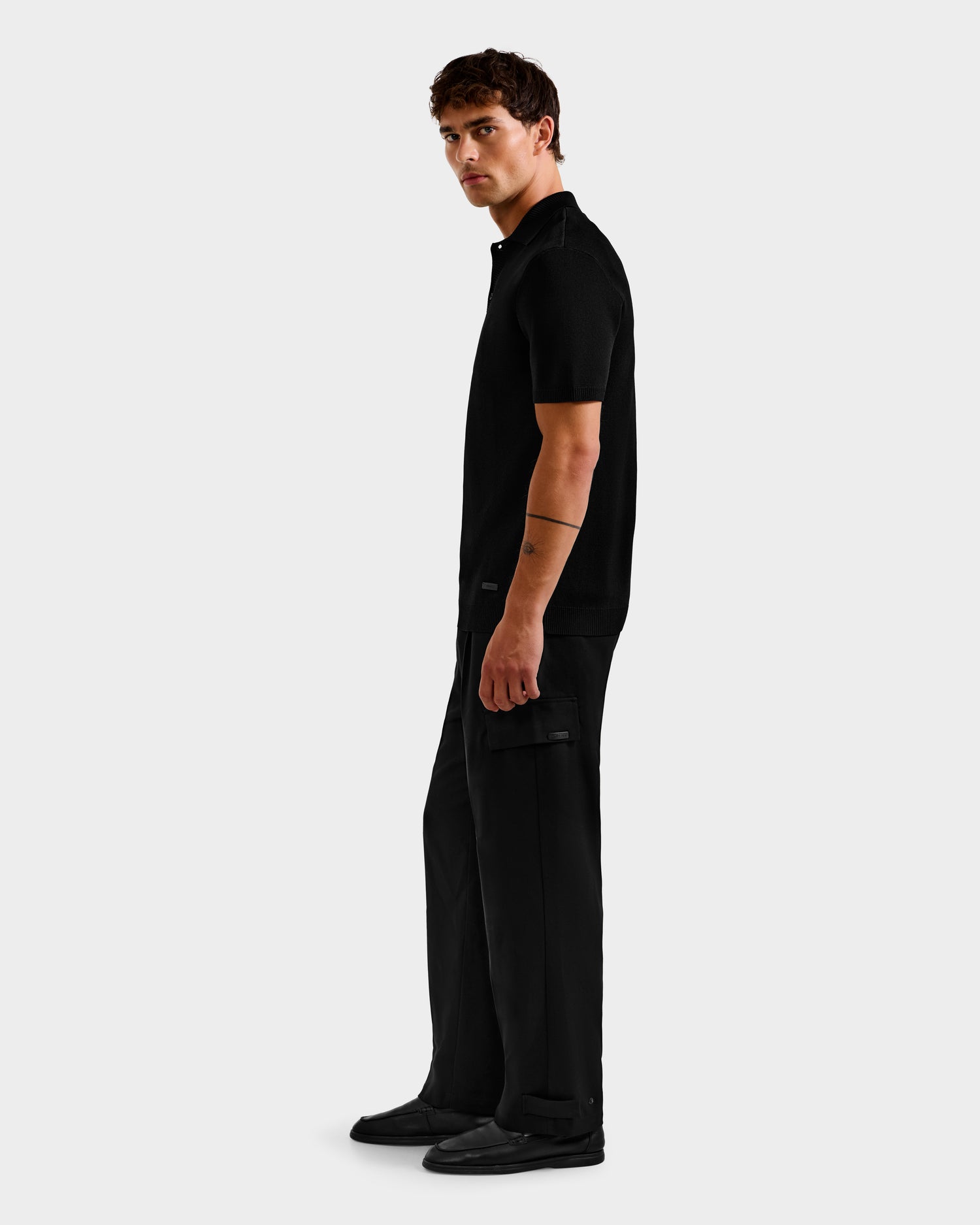 A man stands sideways, looking at the camera, wearing the Rudsak MAISEN - 6524518 Black polo with black cargo pants and shoes against a plain light gray background—a modern essential for men's wardrobes.