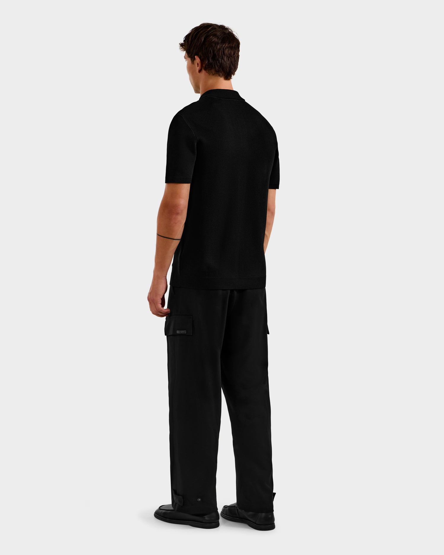A person faces away, wearing Rudsak's MAISEN - 6524518 Black polo and black cargo pants with black shoes, highlighting a modern men's wardrobe staple against a plain light gray background.
