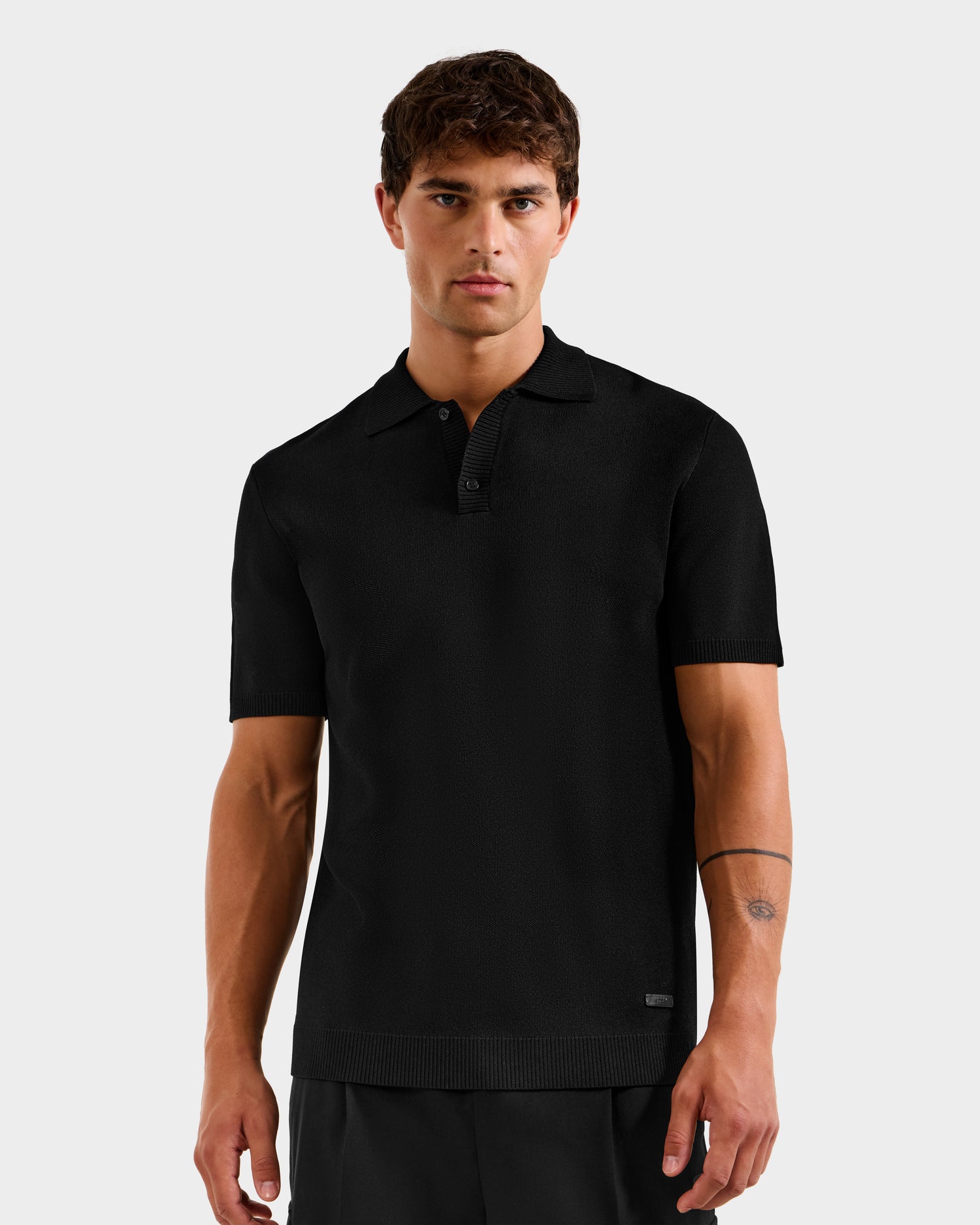 A young man with short brown hair wears the Rudsak MAISEN - 6524518 Black polo and dark pants. He stands against a light gray background, showing a neutral expression and a simple tattoo on his left forearm.