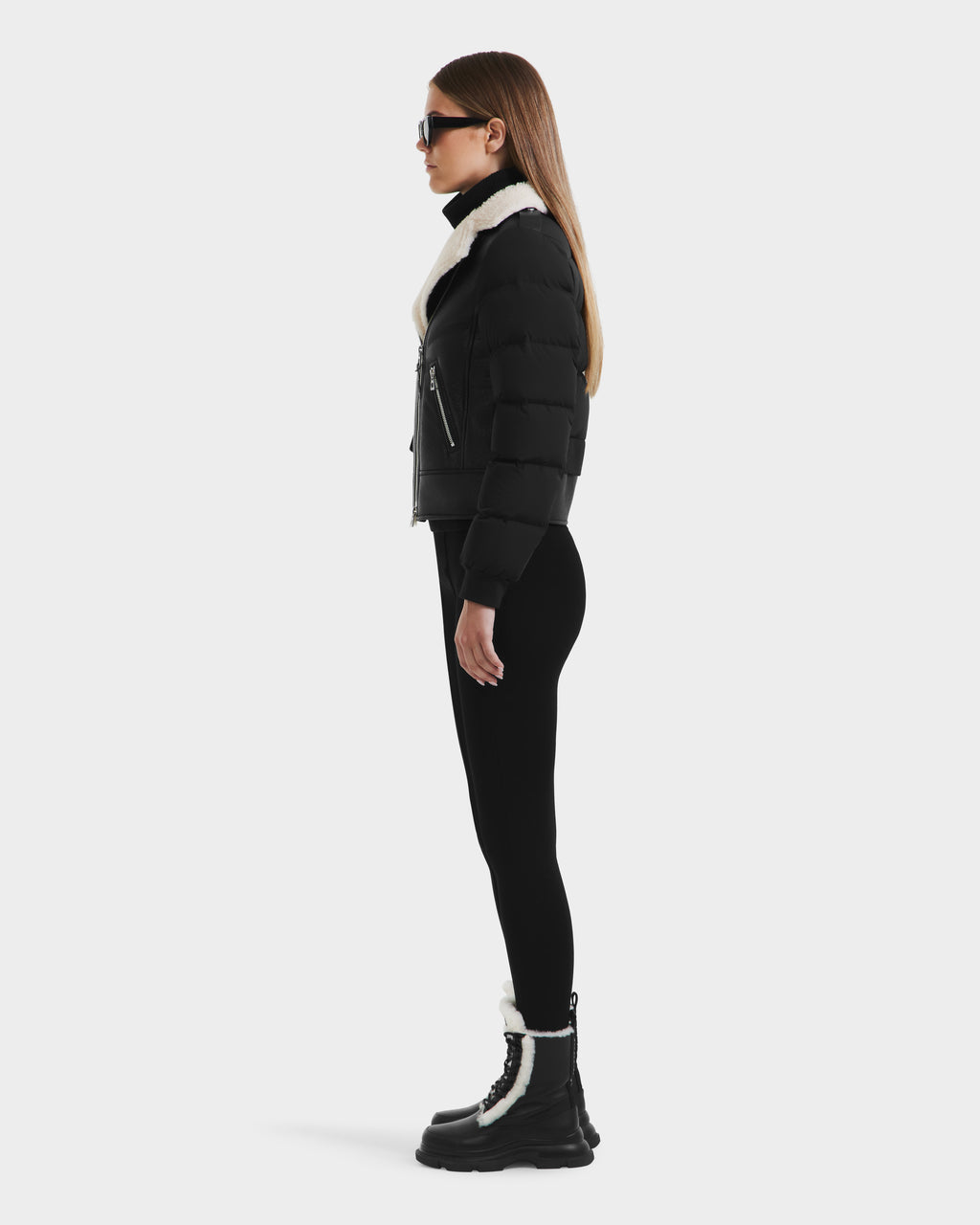 A woman in profile wears Rudsaks MALA - 8125628 Black cropped down jacket with a white shearling collar, styled with black sunglasses, leggings, and black boots with white trim against a plain light background.