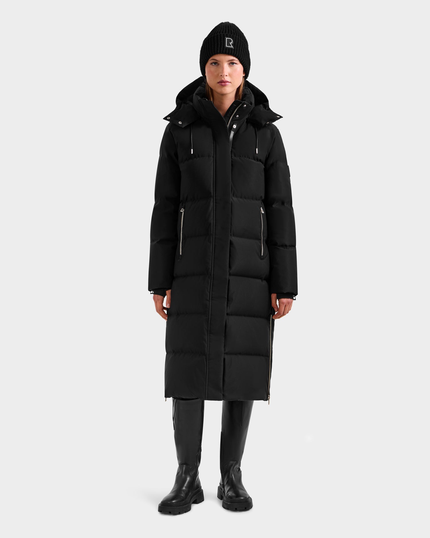 A person stands facing forward against a plain light gray background, wearing the Rudsak MAO H - 8125603 Black women's long down puffer with removable hood, paired with a black beanie, black boots, and black pants.