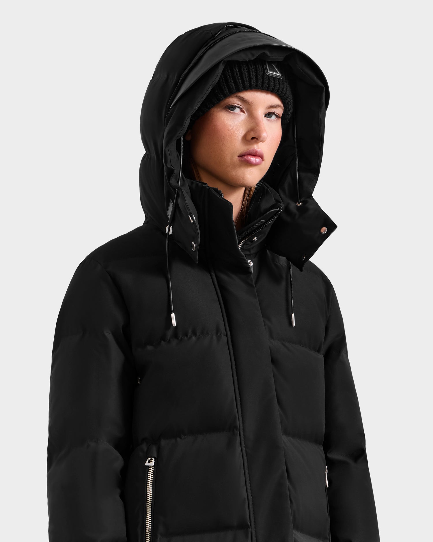 Wearing the Rudsak MAO H - 8125603 Black women’s long down puffer with removable hood and a black knit beanie, she looks slightly to the side against a plain light background.