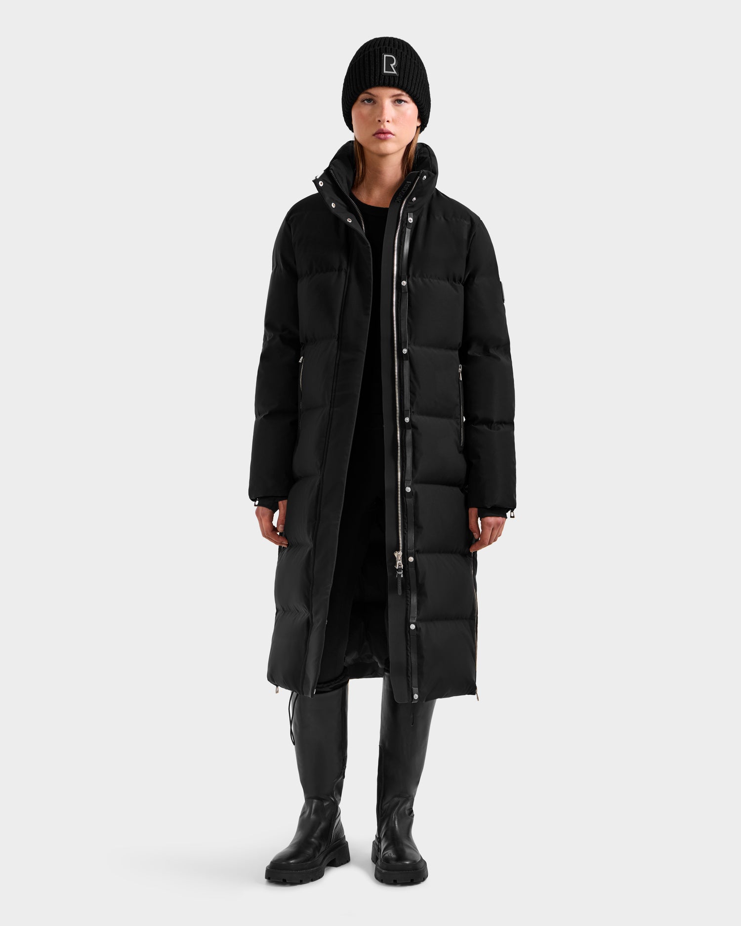 A person stands facing forward, wearing the Rudsak MAO H - 8125603 Black women’s long down puffer with a removable hood, plus a black beanie, dress, boots, and leggings against a plain light gray background.