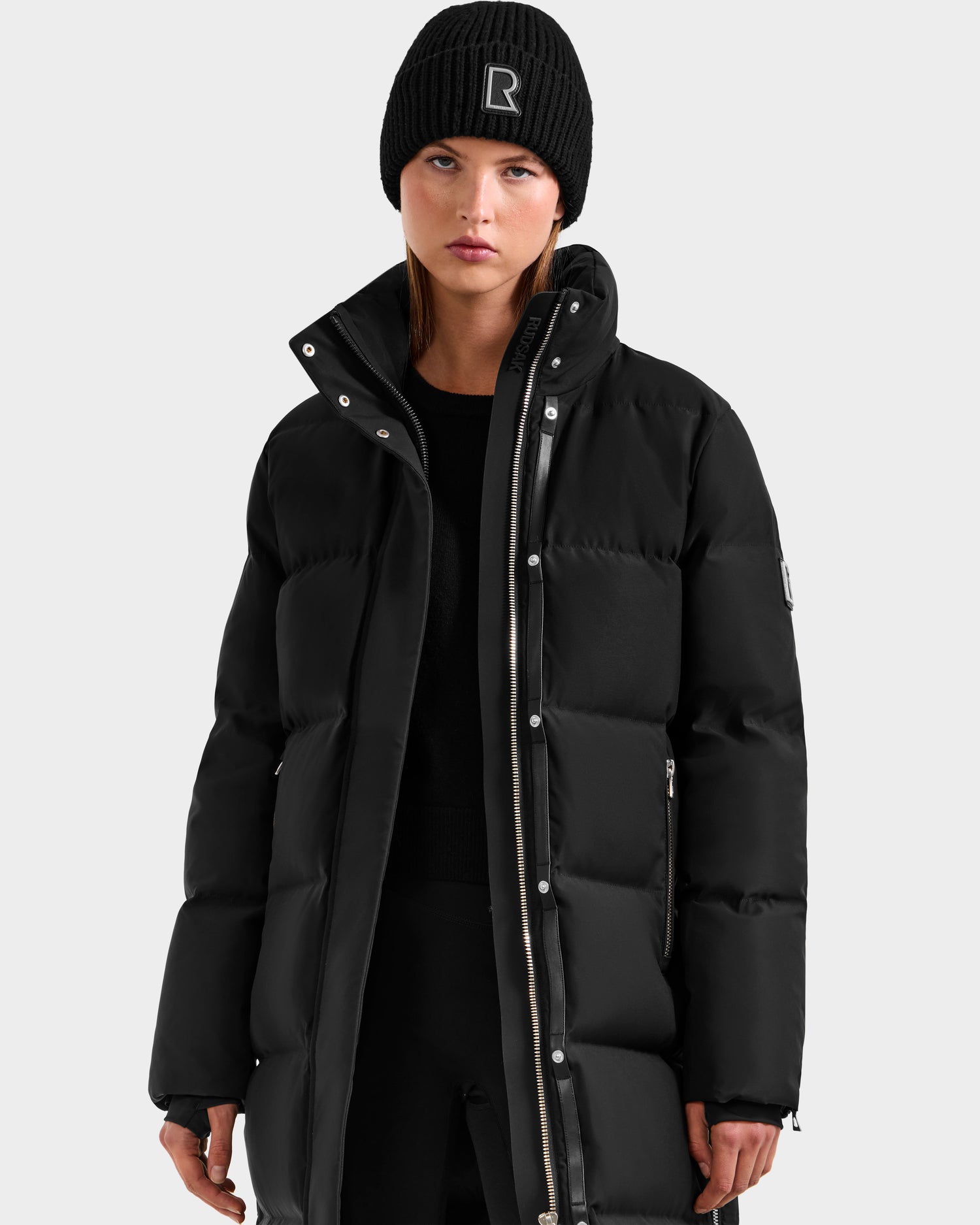 A woman wearing the Rudsak MAO H - 8125603 Black long down puffer jacket and a black beanie with an embroidered R on the front stands against a plain light gray background.