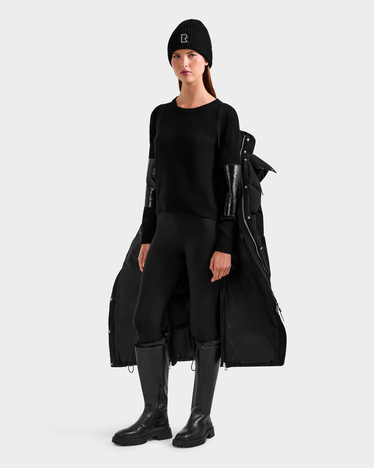 A woman faces forward wearing the Rudsak MAO H - 8125603 Black weather-resistant winter coat with removable hood, black sweater, leggings, tall boots, and a long black coat draped over her shoulders against a light gray background.