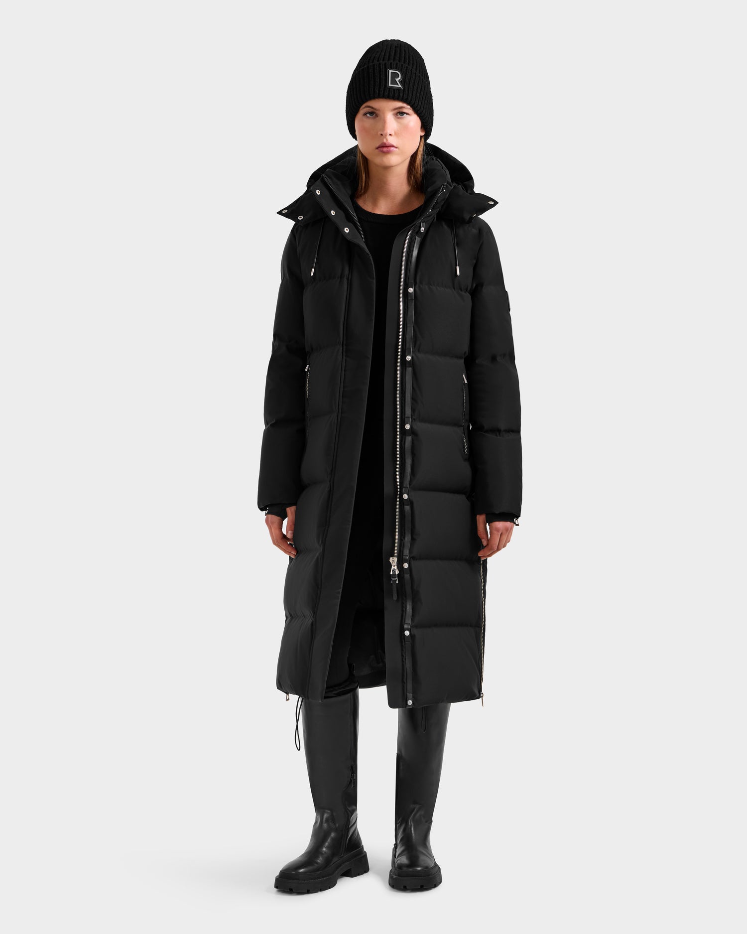 A person wearing the Rudsak MAO H - 8125603 Black women’s long down puffer with a removable hood, plus a black beanie, black dress, and knee-high boots stands facing forward against a plain light gray background.