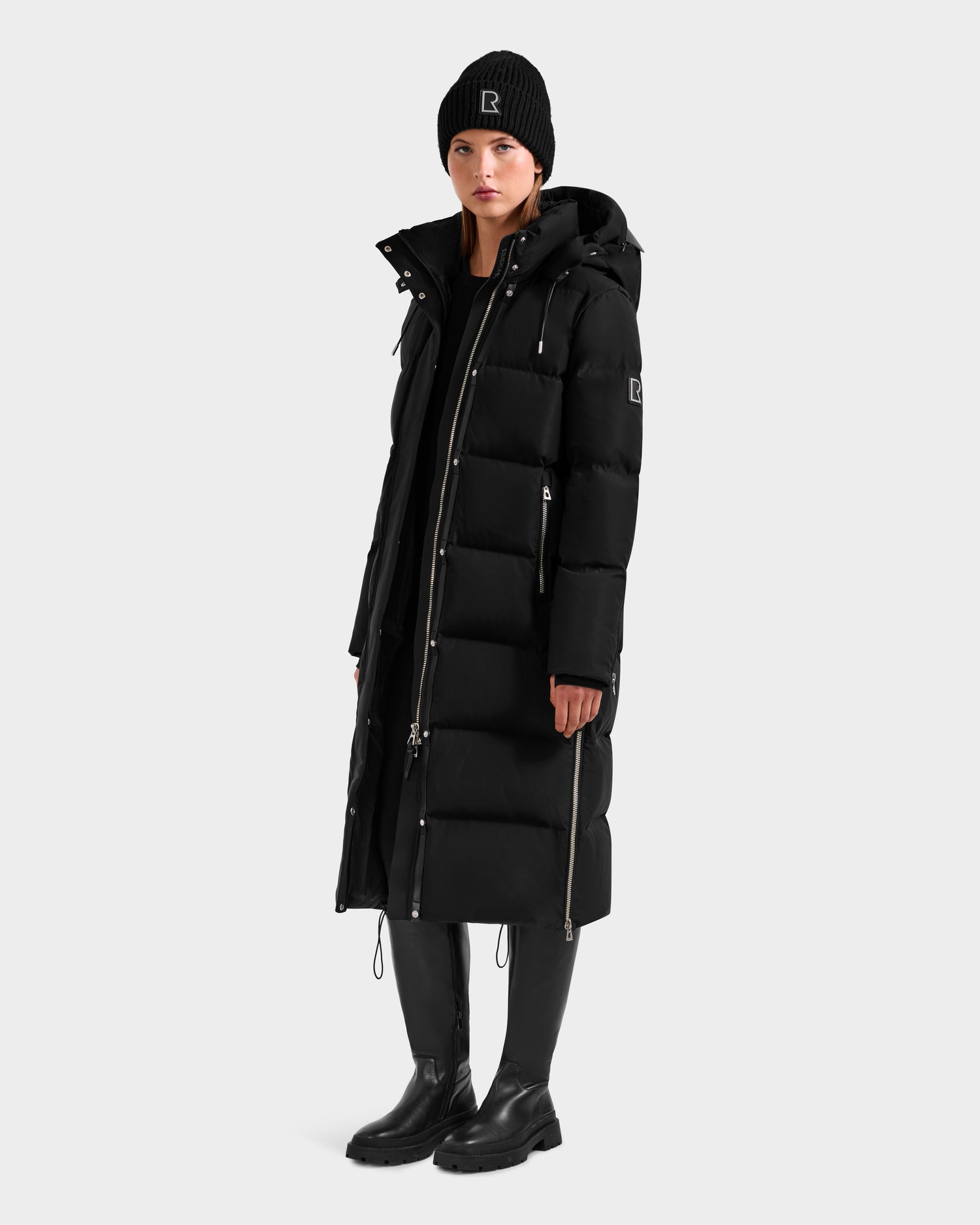 A woman wearing the Rudsak MAO H - 8125603 Black women's long down puffer with a removable hood, black beanie, pants, and boots stands against a light gray background. The weather-resistant coat features front zippers and extends below the knees.
