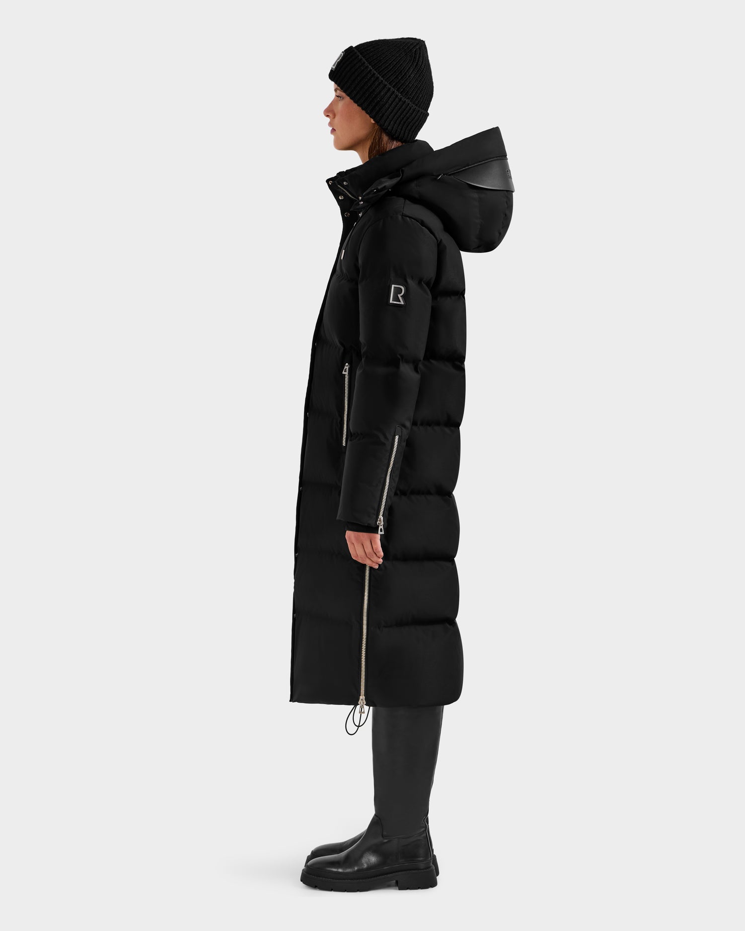 A person in profile wears the Rudsak MAO H - 8125603 Black women's long down puffer with a removable hood, black boots, and a black knit beanie, standing against a plain light gray background.
