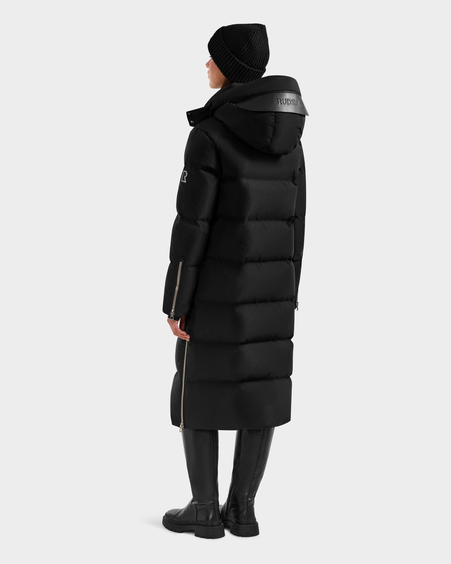 A person in the Rudsak MAO H - 8125603 Black women's long down puffer with removable hood, black knit hat, and knee-high boots stands facing away, head slightly turned, against a plain light background.