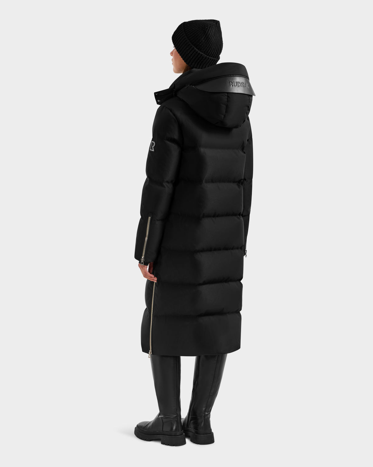 A person in the Rudsak MAO H - 8125603 Black women's long down puffer with removable hood, black knit hat, and knee-high boots stands facing away, head slightly turned, against a plain light background.