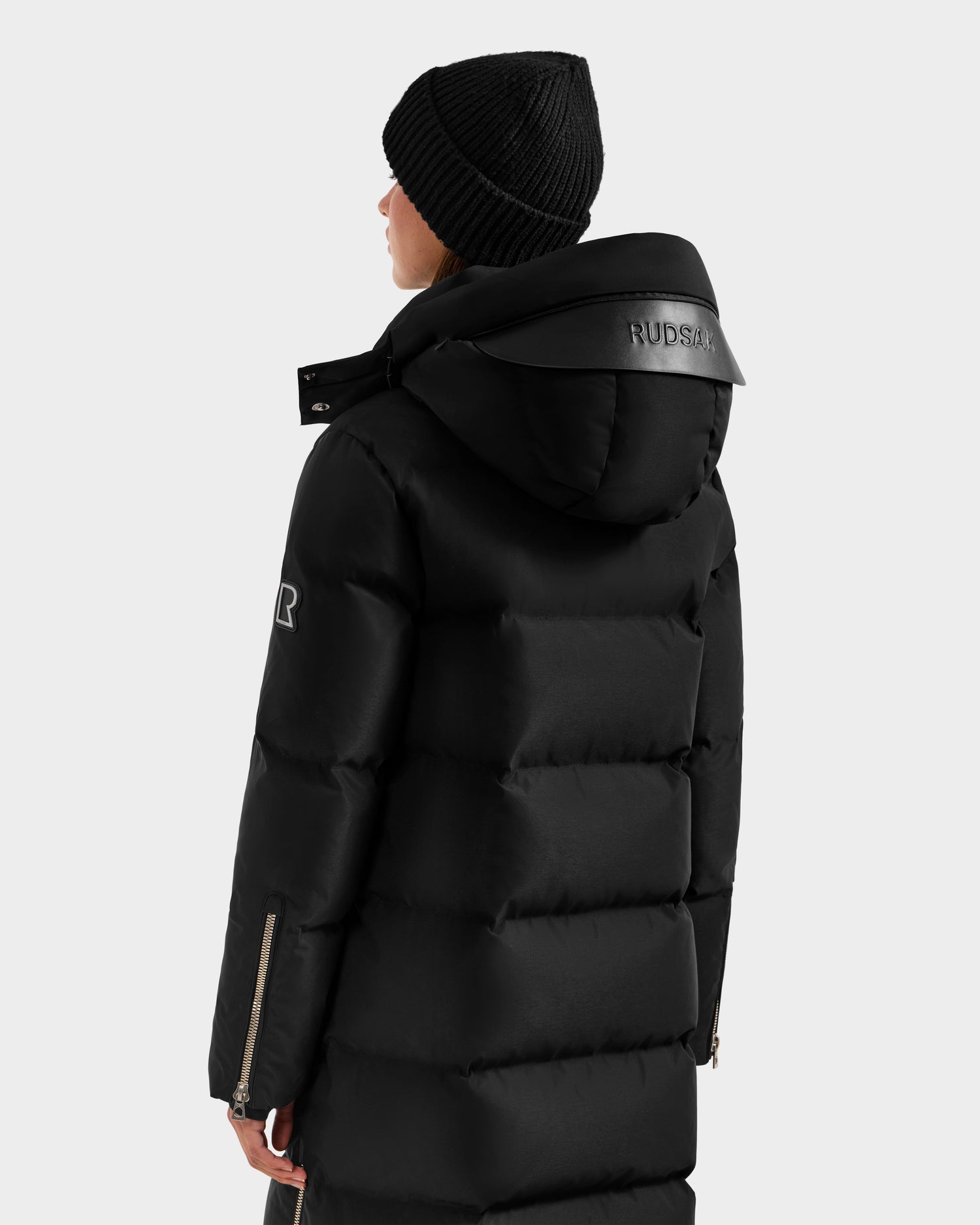 A person seen from behind wears the Rudsak MAO H - 8125603 Black women’s long down puffer with hood and a black knit beanie. The weather-resistant coat features “RUDSAK” branding on the hood and a zippered sleeve detail.