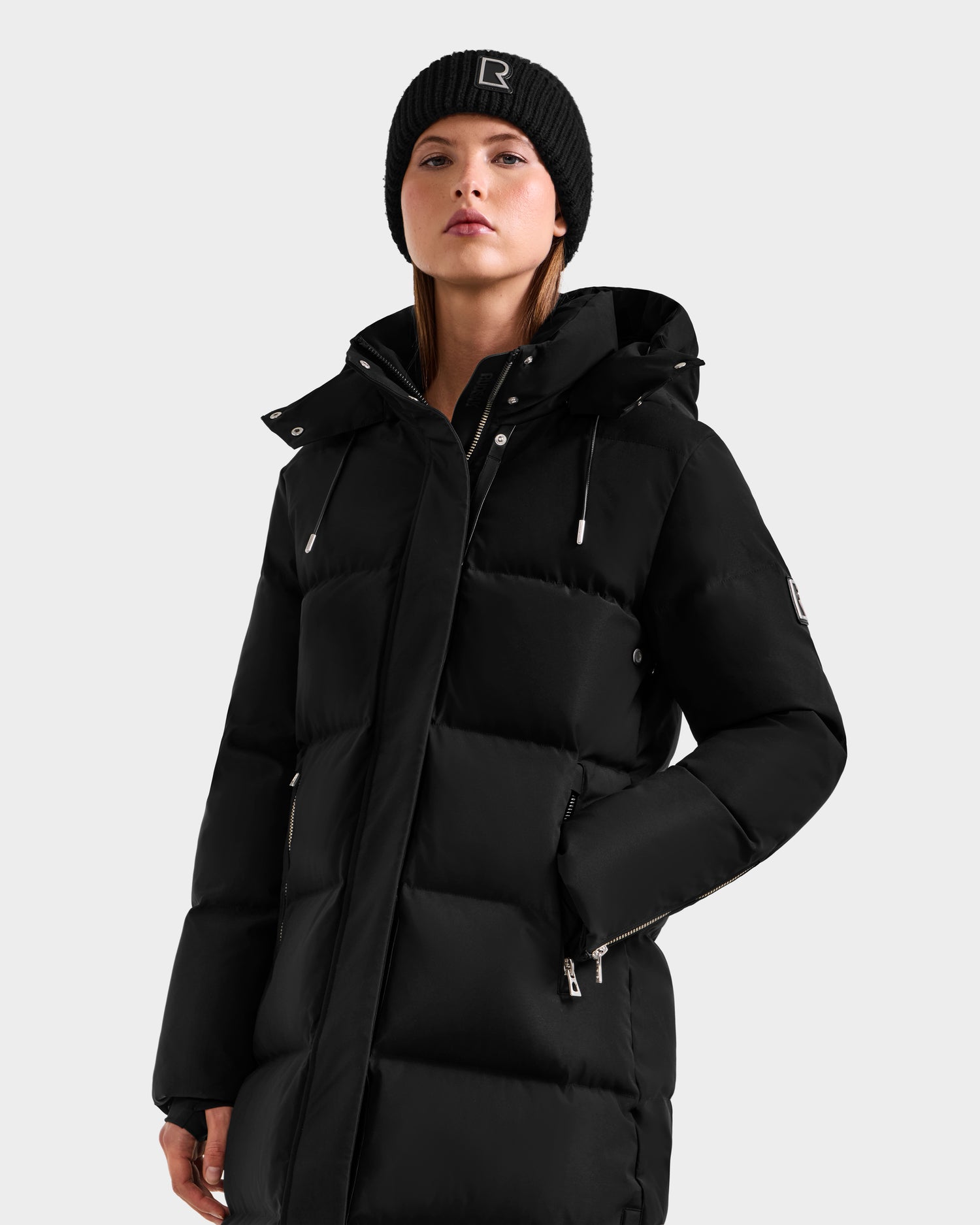 A woman confidently poses in the Rudsak MAO H - 8125603 Black winter coat—weather-resistant, zippered pockets, removable hood—and a black knit beanie with an "R.