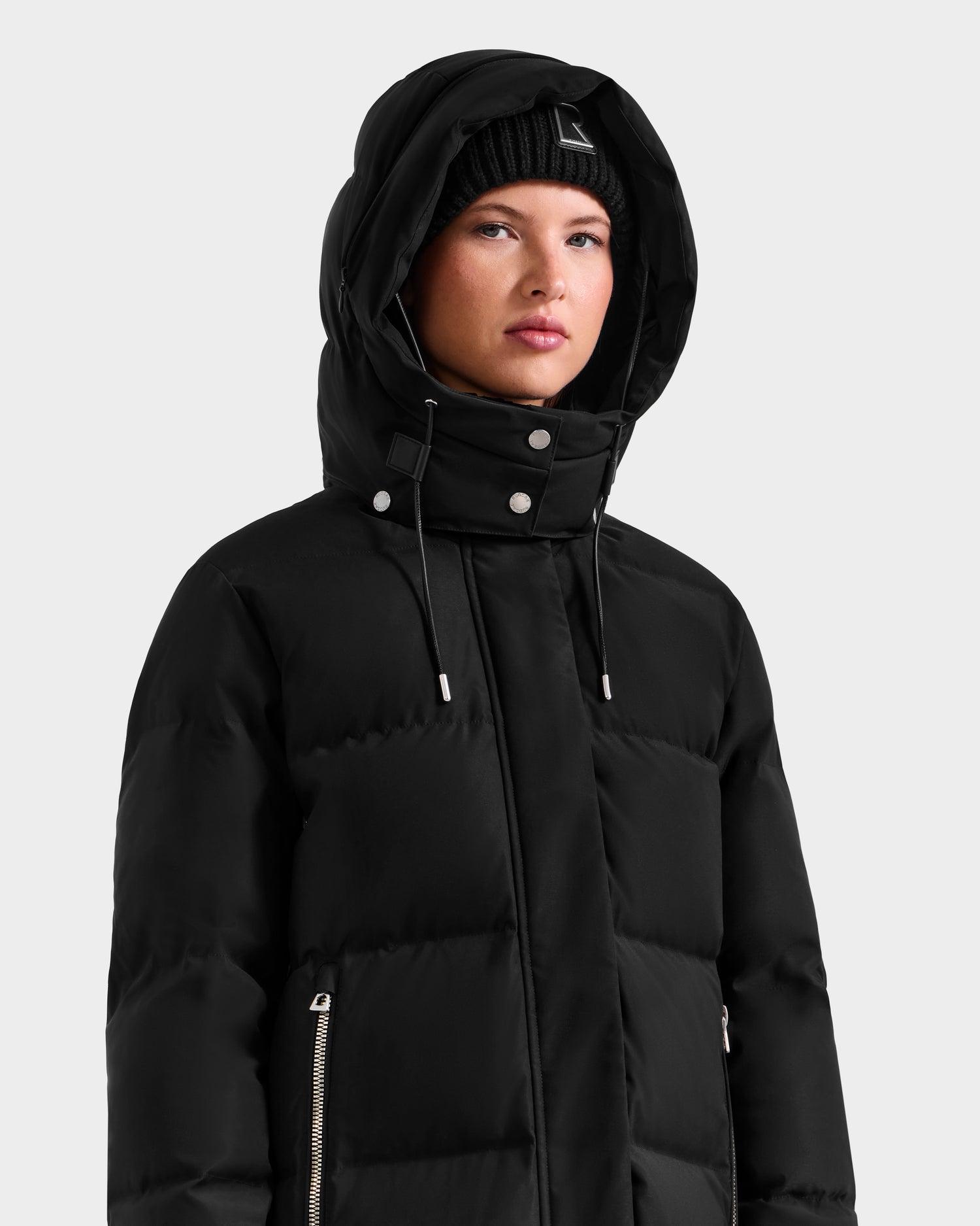 Wearing the Rudsak MAO H - 8125603 Black puffer jacket with hood and a black knit beanie, a person stands against a plain light gray background, looking forward with a neutral expression.