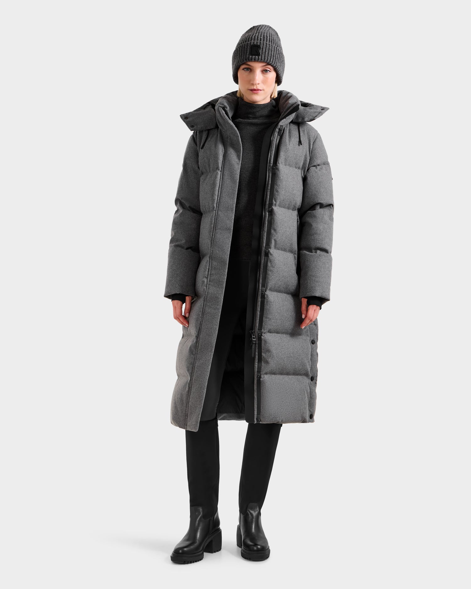 A person stands facing forward in the Rudsak MAO W - 8125568 Heather Charcoal long puffer coat with a wool-blend shell, black pants, black boots, and a gray knit beanie against a plain light background.