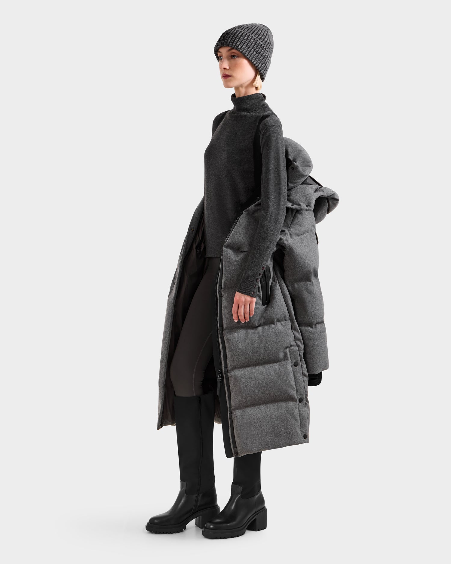 A person wearing a dark turtleneck, leggings, tall black boots, and a gray beanie stands sideways holding the Rudsak MAO W - 8125568 Heather Charcoal wool-blend shell puffer coat over one arm against a plain light background.