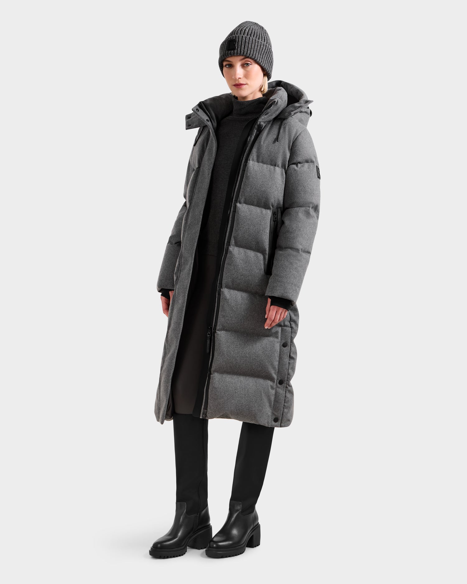 A person wears the Rudsak MAO W - 8125568 Heather Charcoal long puffer coat with a wool-blend shell, black pants, boots, a black sweater, and a gray knit beanie against a plain light background.