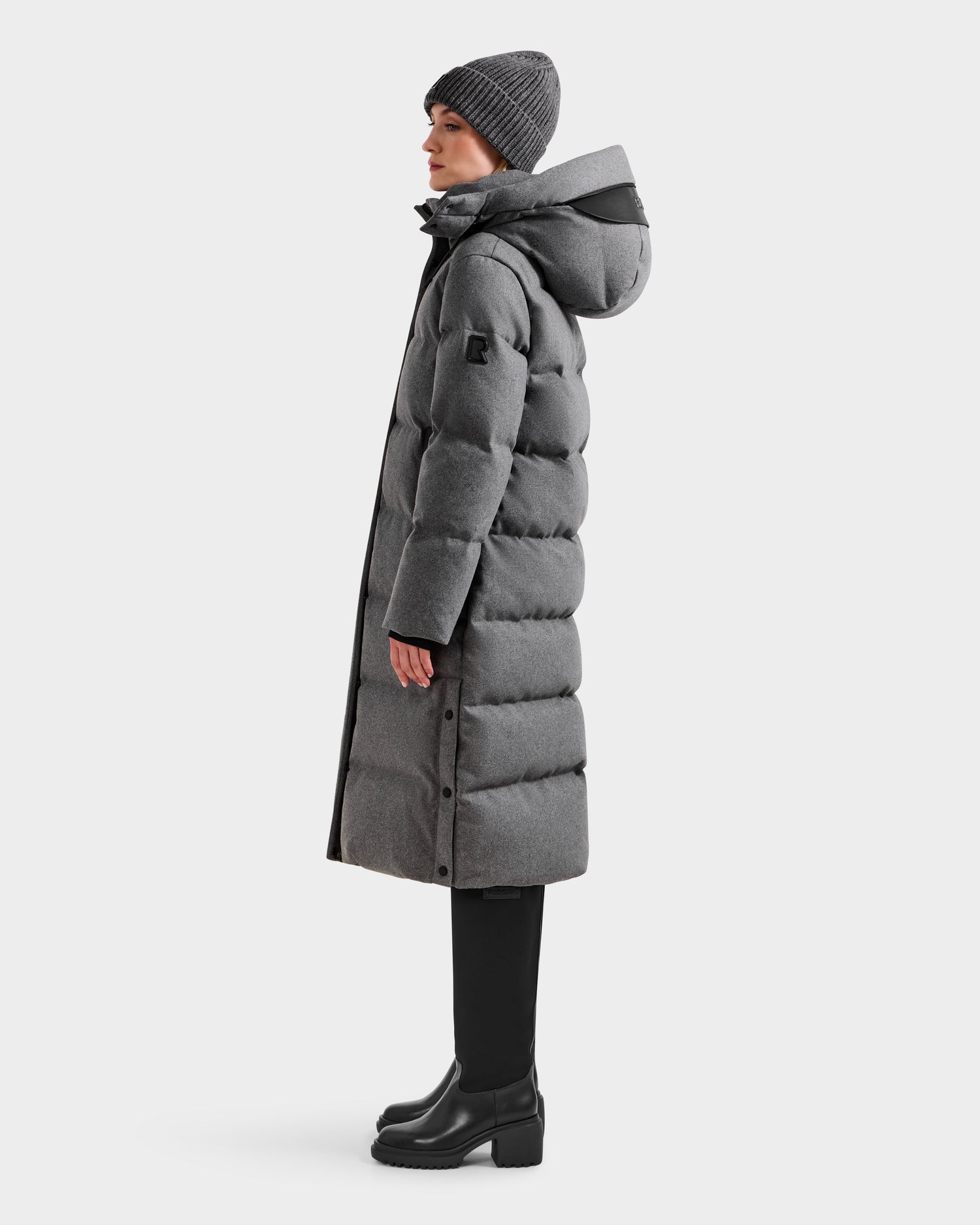 A person in profile wears the Rudsak MAO W - 8125568 Heather Charcoal puffer coat with a hood, styled with a gray beanie, black pants, and black heeled boots against a plain white background.