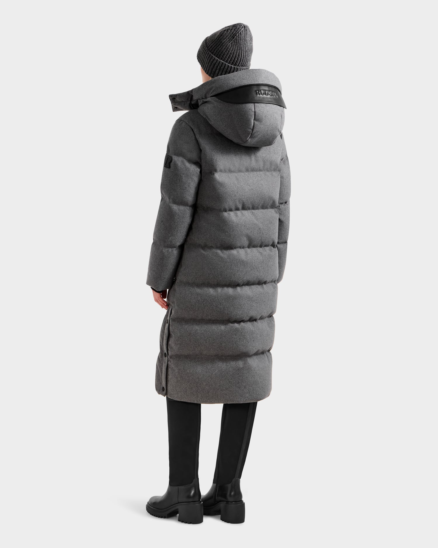 A person wearing the Rudsak MAO W - 8125568 Heather Charcoal puffer coat with a hood, black pants, black boots, and a gray knit beanie stands facing away from the camera against a plain background.