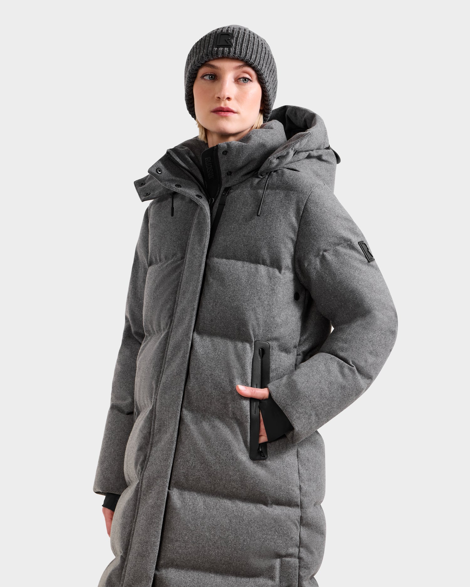 A person wearing the Rudsak MAO W - 8125568 Heather Charcoal long puffer coat with a wool-blend shell and hood, plus a gray knit beanie, stands against a light background, looking to the side with one hand in their pocket.